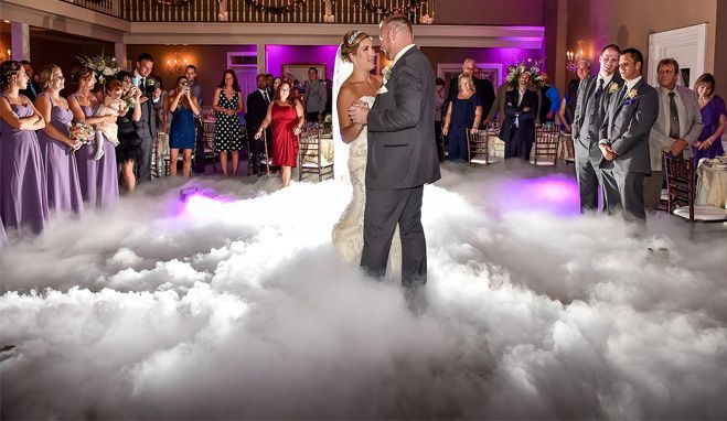 Bride and groom dance on a cloud of fog at a wedding reception; guests watch.