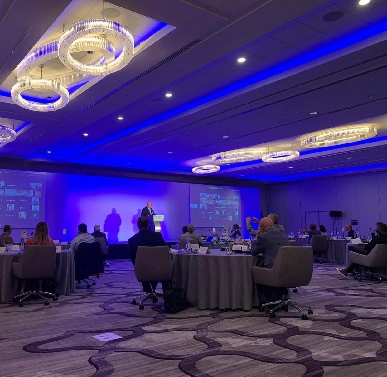 A group of people are sitting at tables in a large room with blue lights.