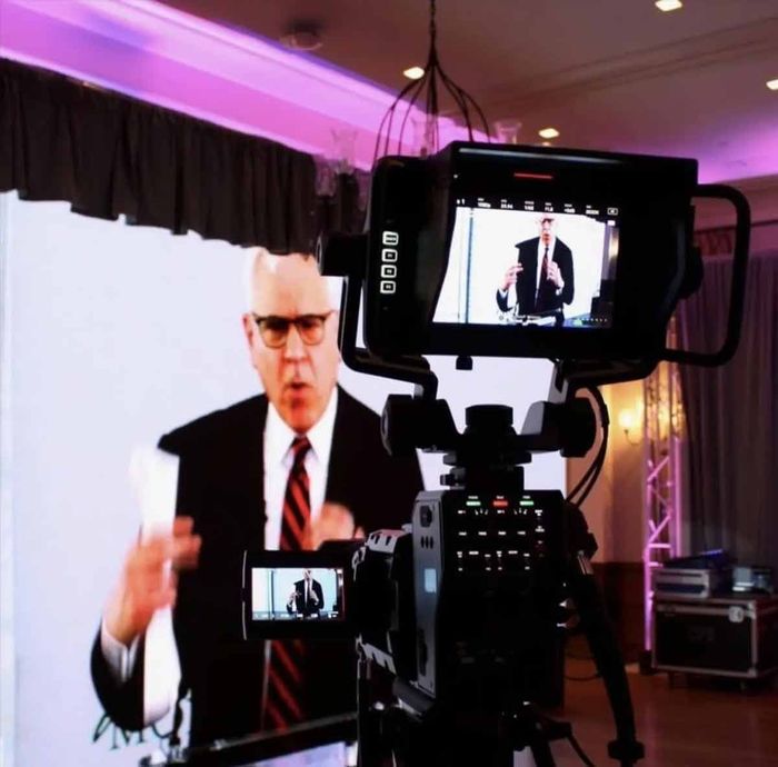 A professional video camera records a person in a suit and tie speaking in front of a white backdrop in a lit room.