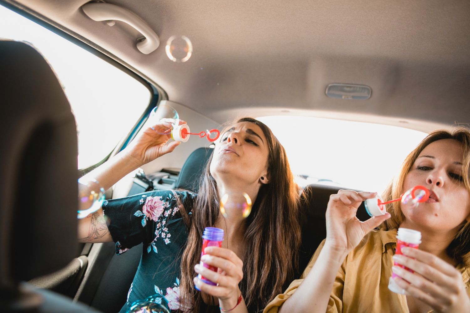 Two women are blowing soap bubbles in the back seat of a car.