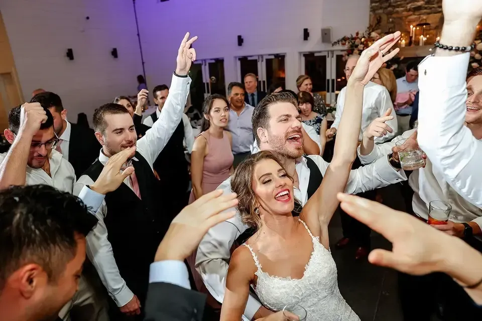 Wedding reception with dancing guests, including a bride and groom raising their arms with joy.