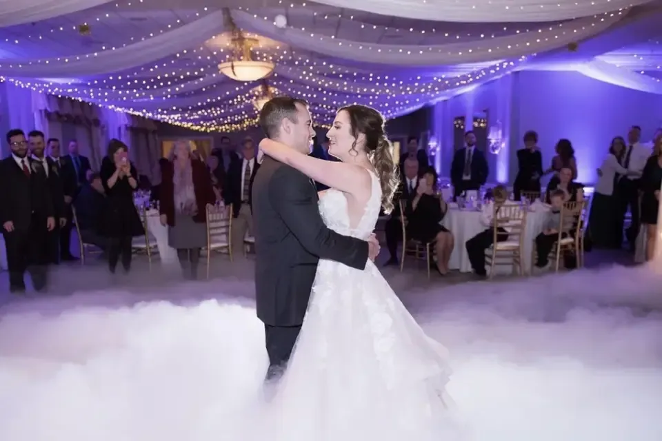 Couple dancing on a fog-covered dance floor during a wedding reception, surrounded by guests, with blue and white lighting.
