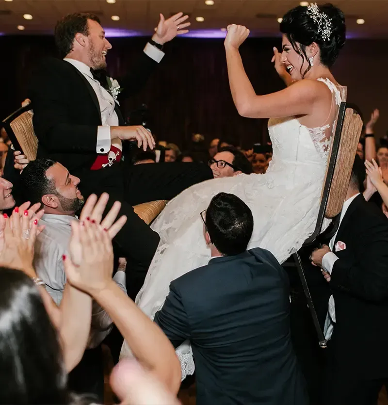 Couple in wedding attire being lifted on chairs, celebrating. Guests clap, smiling in a ballroom setting.