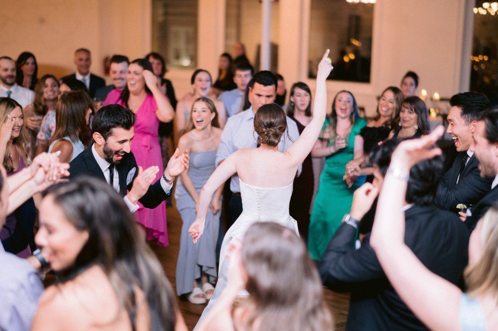 Bride dancing with arms raised, surrounded by cheering guests at a wedding reception.