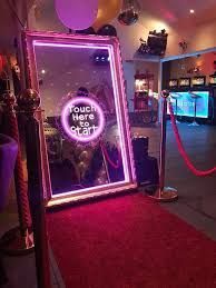 A magic mirror photo booth with pink neon, on a red carpet with stanchions, set in a party location.