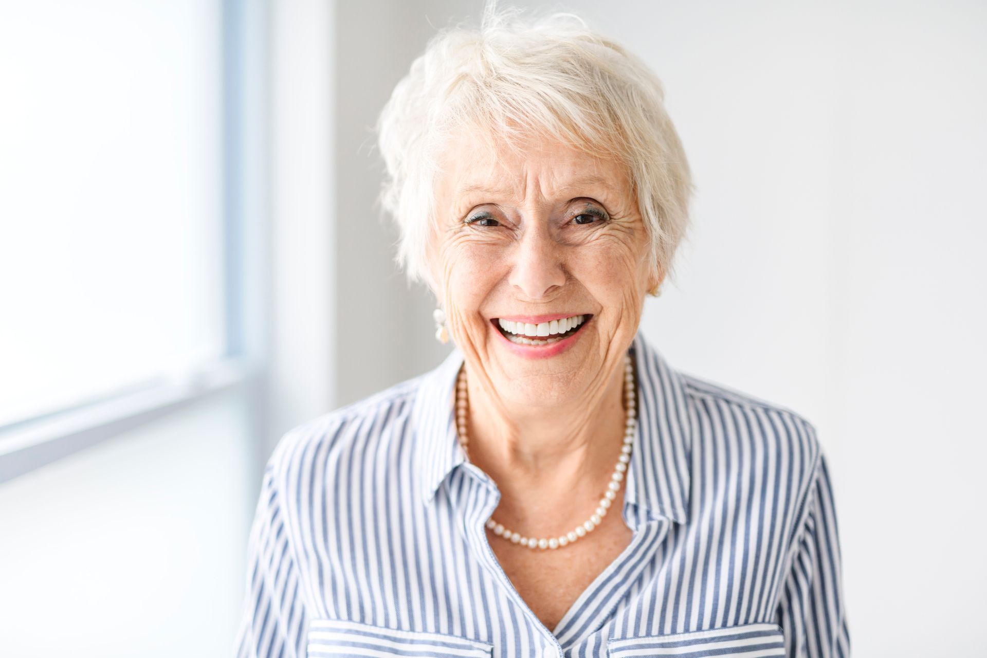 An elderly woman wearing a striped shirt and a pearl necklace is smiling for the camera.