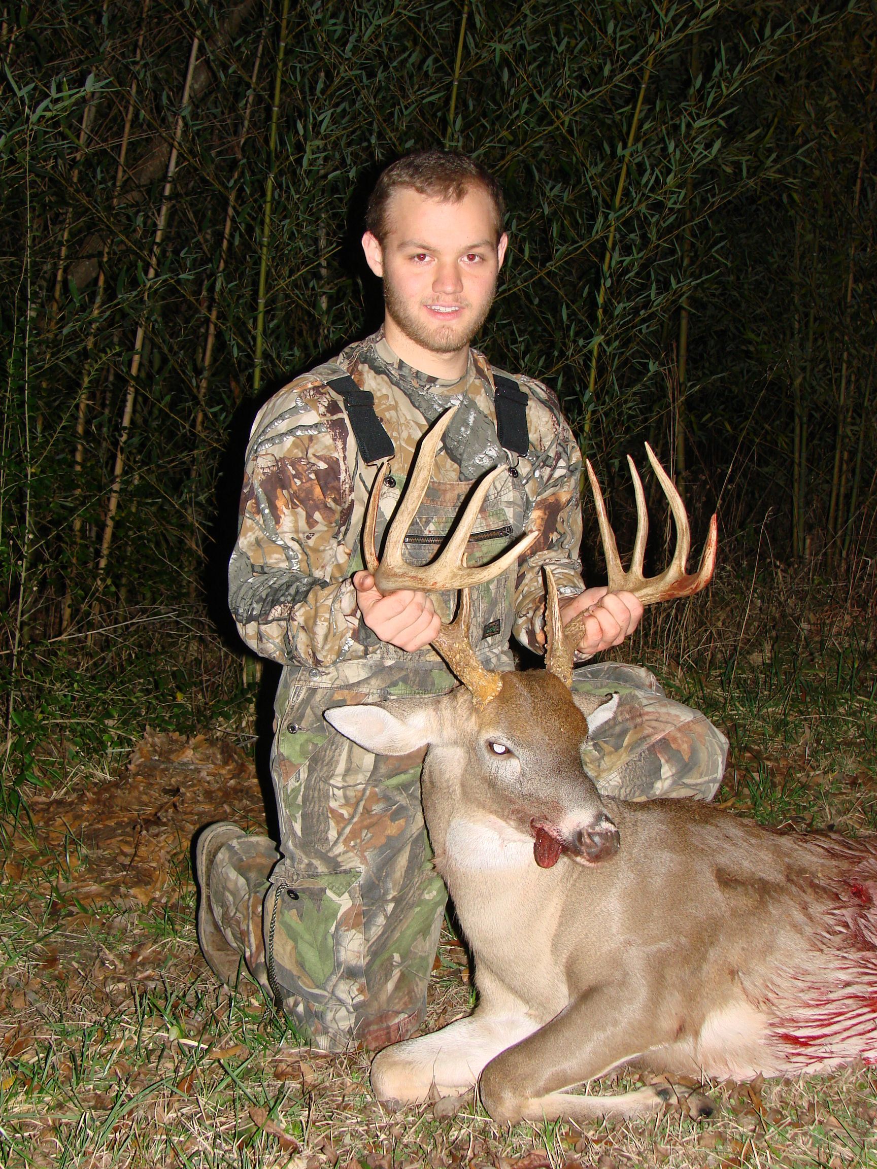 A man in camouflage kneels beside a dead buck he has hunted. He holds the antlers and looks at the camera. The deer has blood on its mouth.