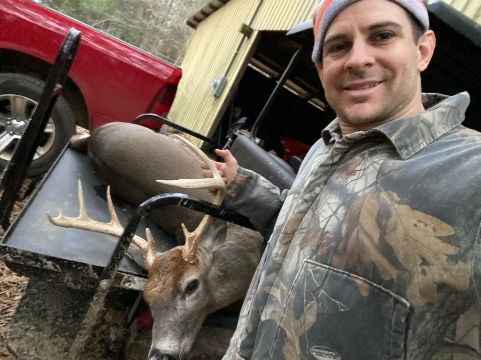 Man in camouflage holding a deer with antlers next to a red truck and a shed.