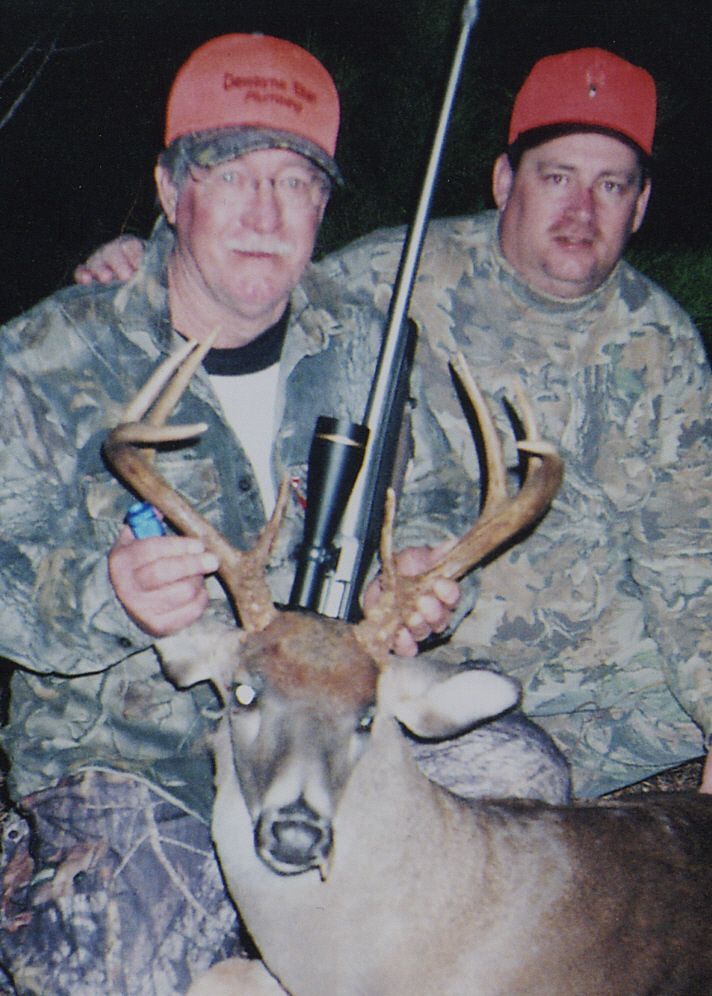 Two men in camo pose with a deer they hunted, holding its antlers and a rifle. They wear orange hats and smile.