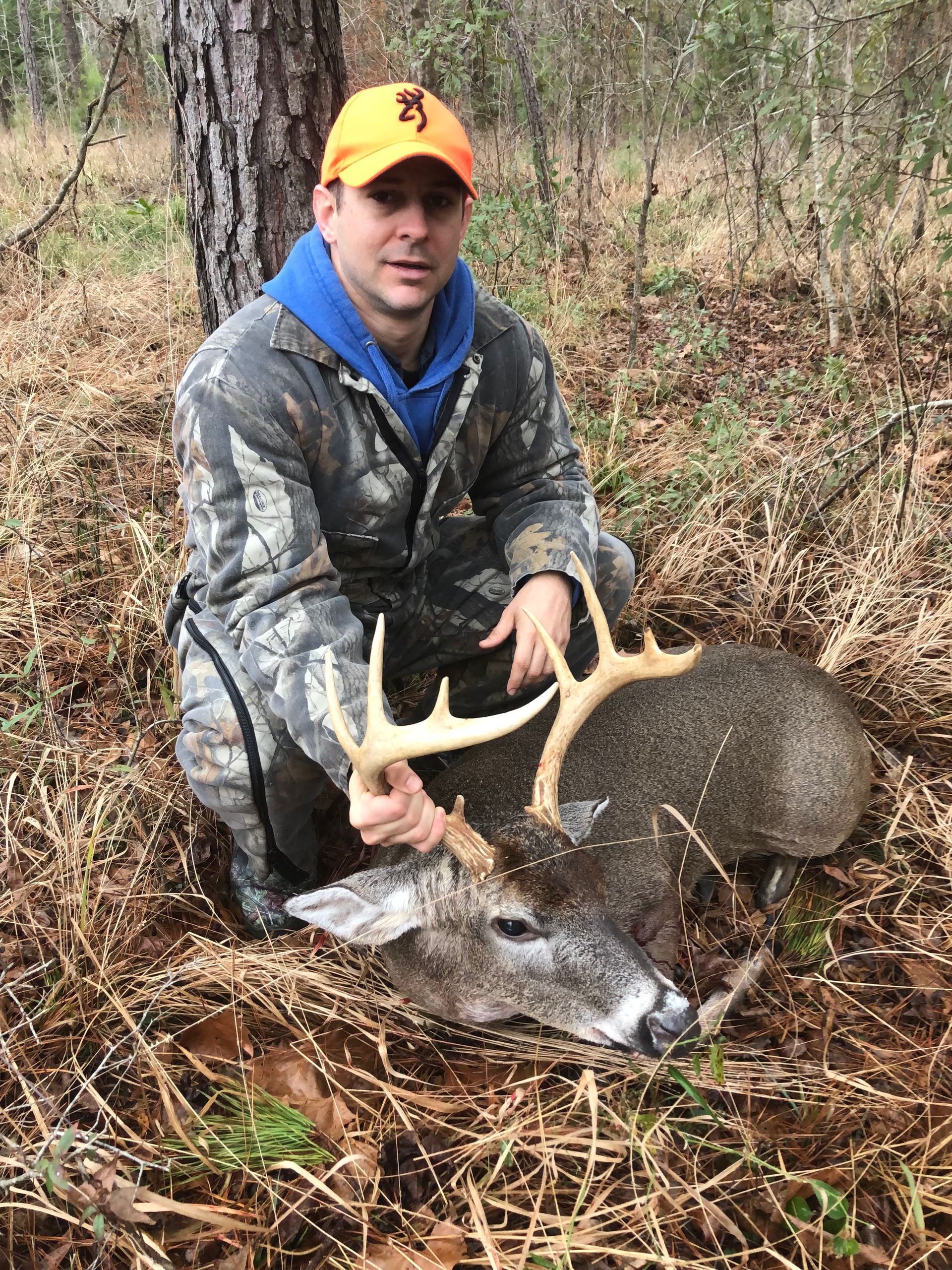 A man kneels next to a harvested deer in a wooded area. He wears camouflage and holds the deer's antlers, smiling.