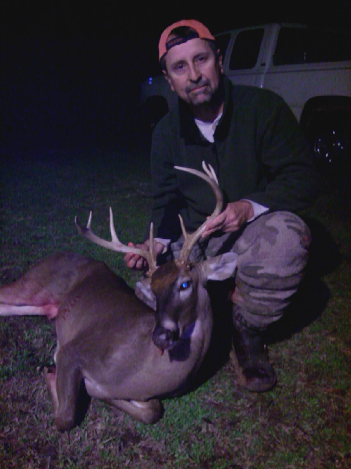 Man in hunting gear kneels beside a buck he harvested. He wears a cap, green jacket, and camo pants. The scene is at night.
