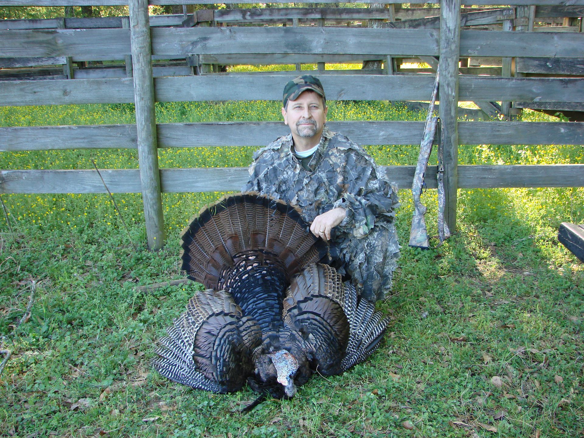 A man in camouflage kneels next to a dead turkey with its feathers fanned out, in front of a wooden fence.