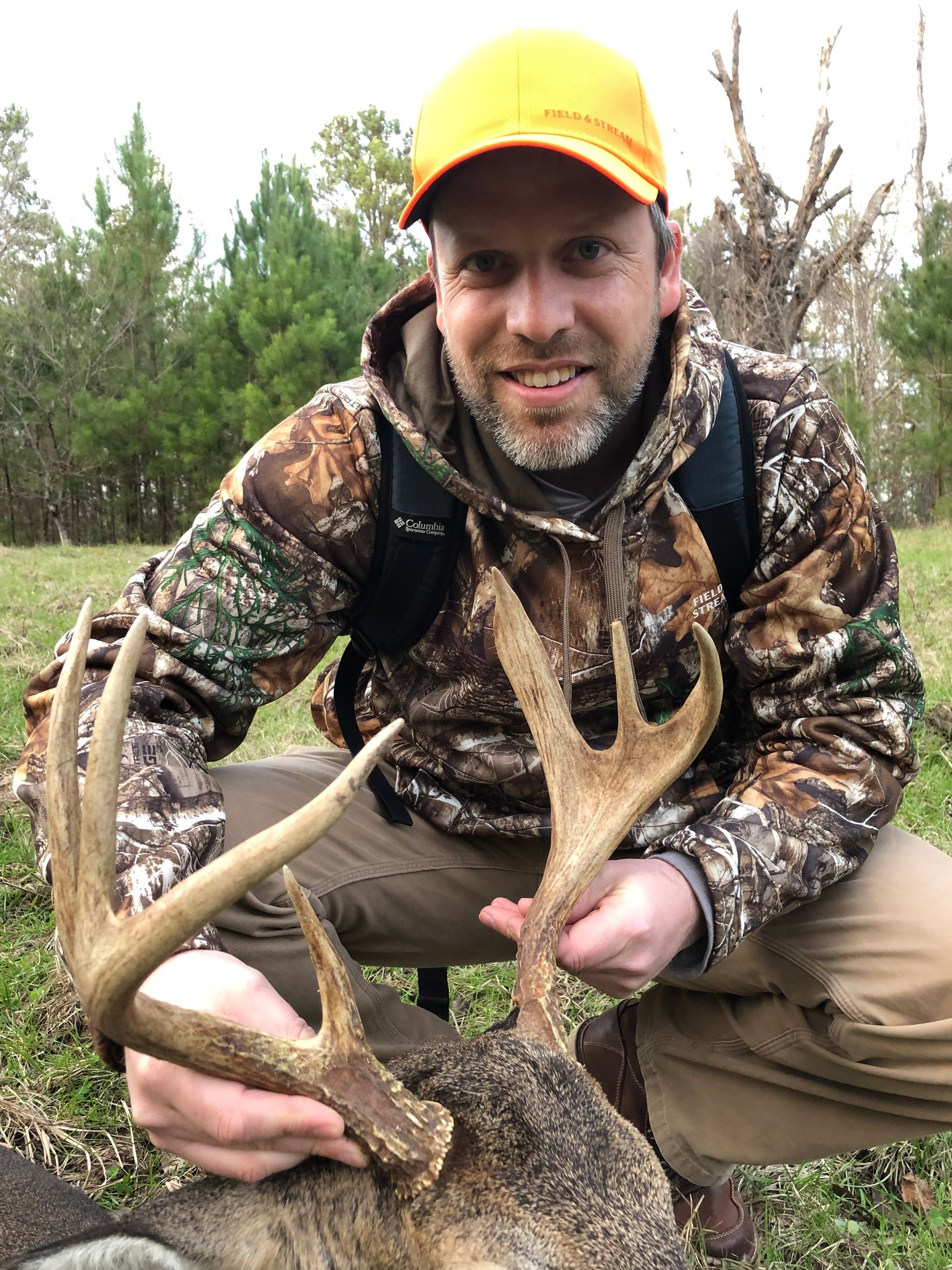Man in camo hunting gear kneels, holding deer antlers; orange cap; outdoors.