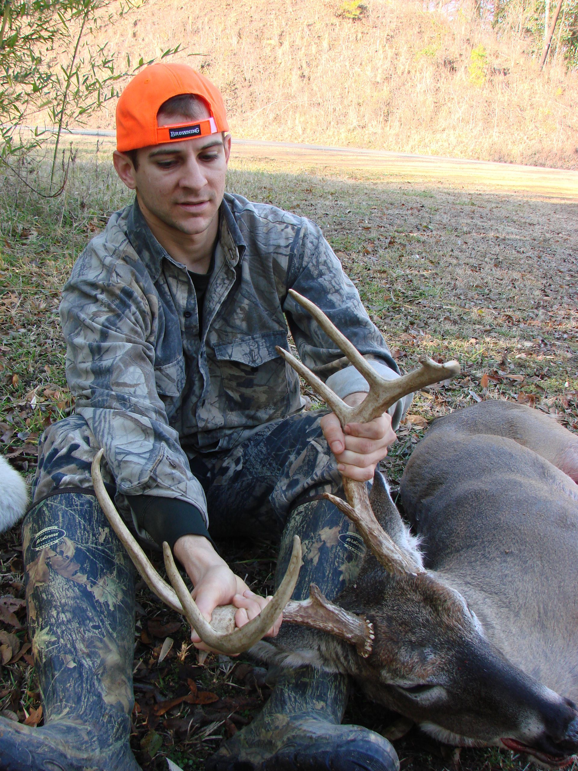 Hunter in camouflage and orange hat, seated and holding deer antlers. Dead deer is lying on the ground.