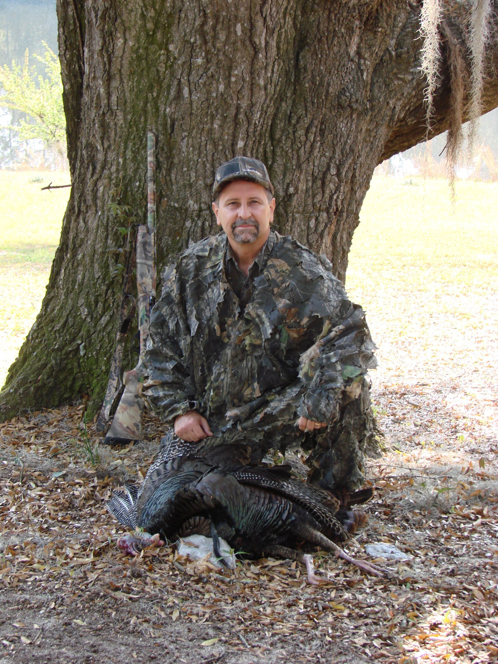 Man in camouflage kneeling beside a harvested turkey under a large tree. He is smiling, and a rifle leans against the tree.
