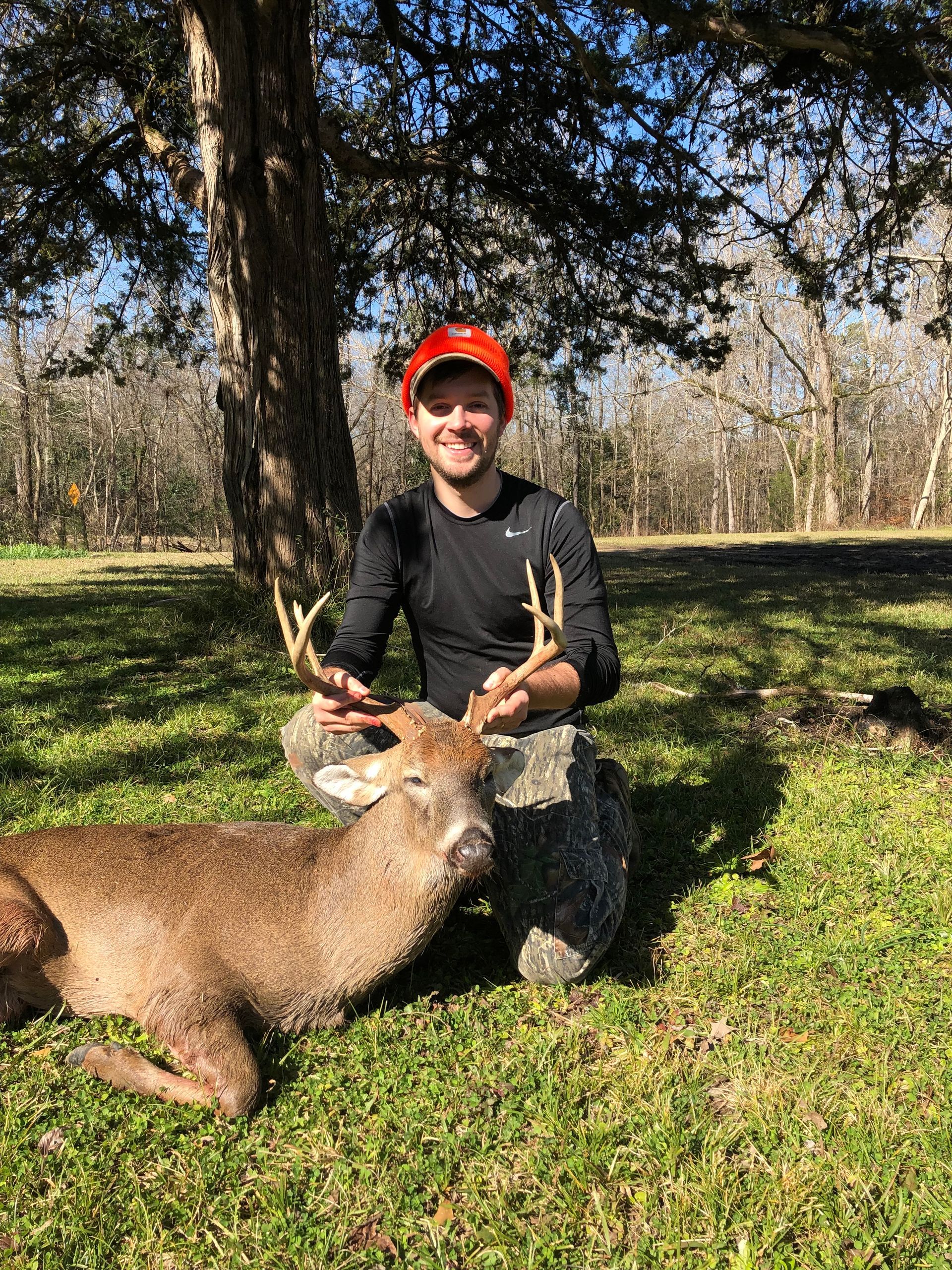 A man kneels beside a deer he has hunted, smiling. The man wears an orange hat and black shirt; the deer is on green grass in a wooded area.