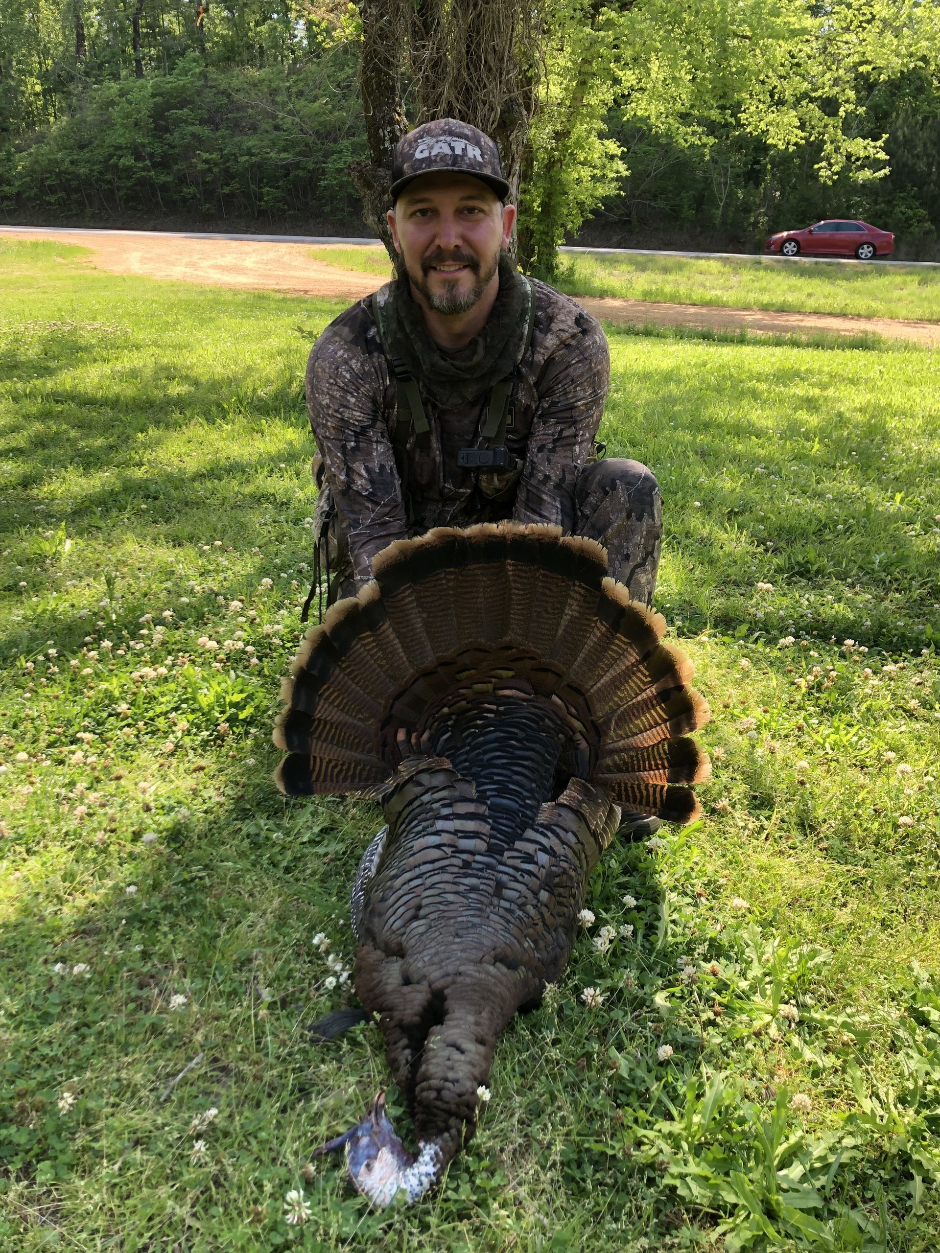 Man in camo kneeling with a harvested turkey, its tail feathers fanned out in a grassy area.