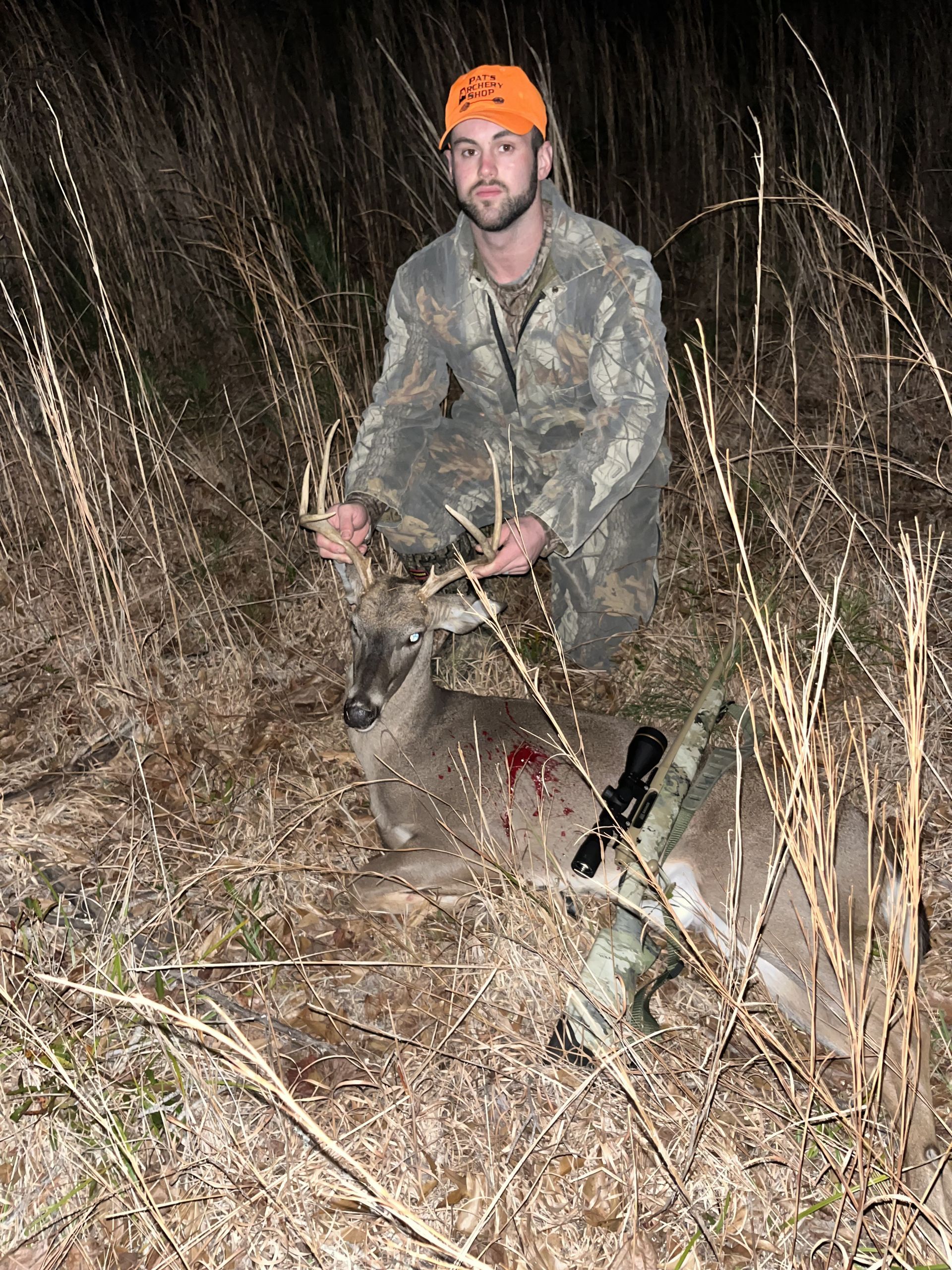 A man in camouflage and an orange hat kneels beside a harvested deer in tall grass at night.