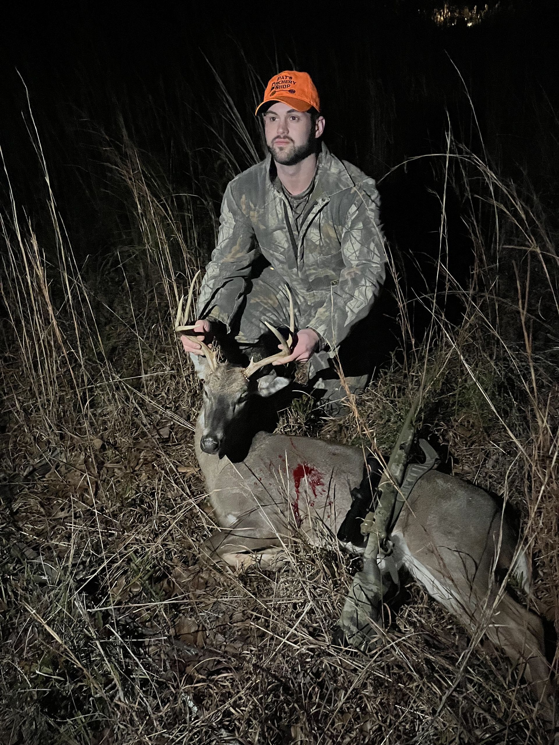 Hunter in camouflage kneels beside a dead deer in a field at night. He wears an orange hat and holds the deer's antlers.
