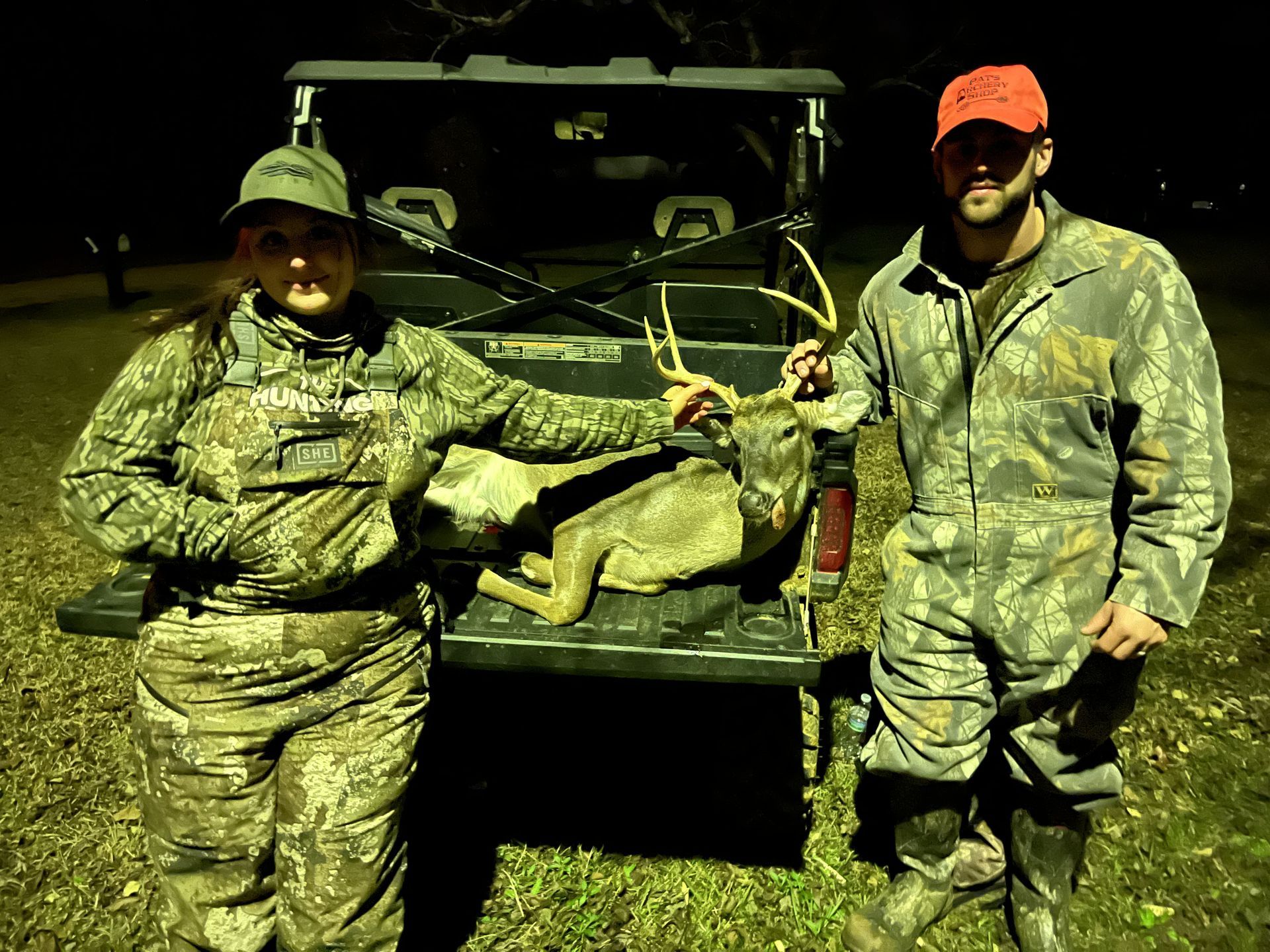 A woman and a man in hunting gear pose with a deer in the bed of a truck at night.