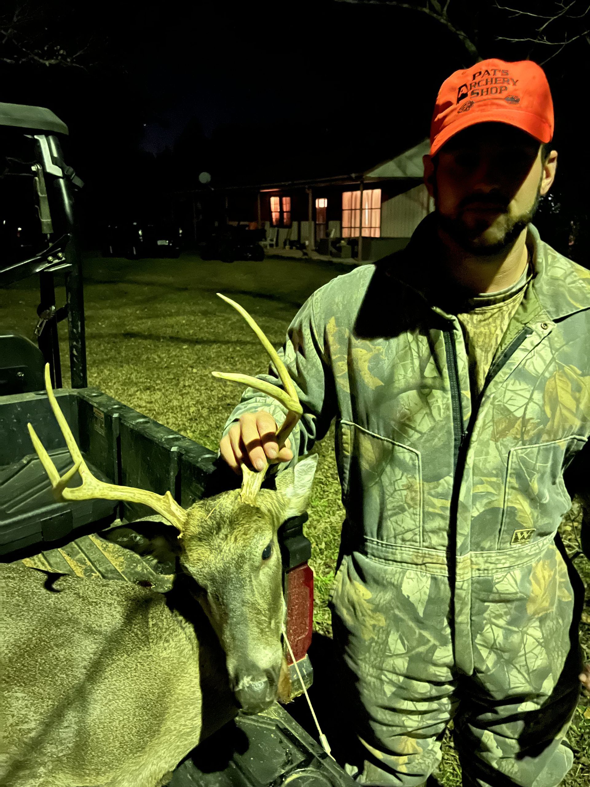 Man in camo and orange cap stands next to a buck with antlers in front of a vehicle and house at night.