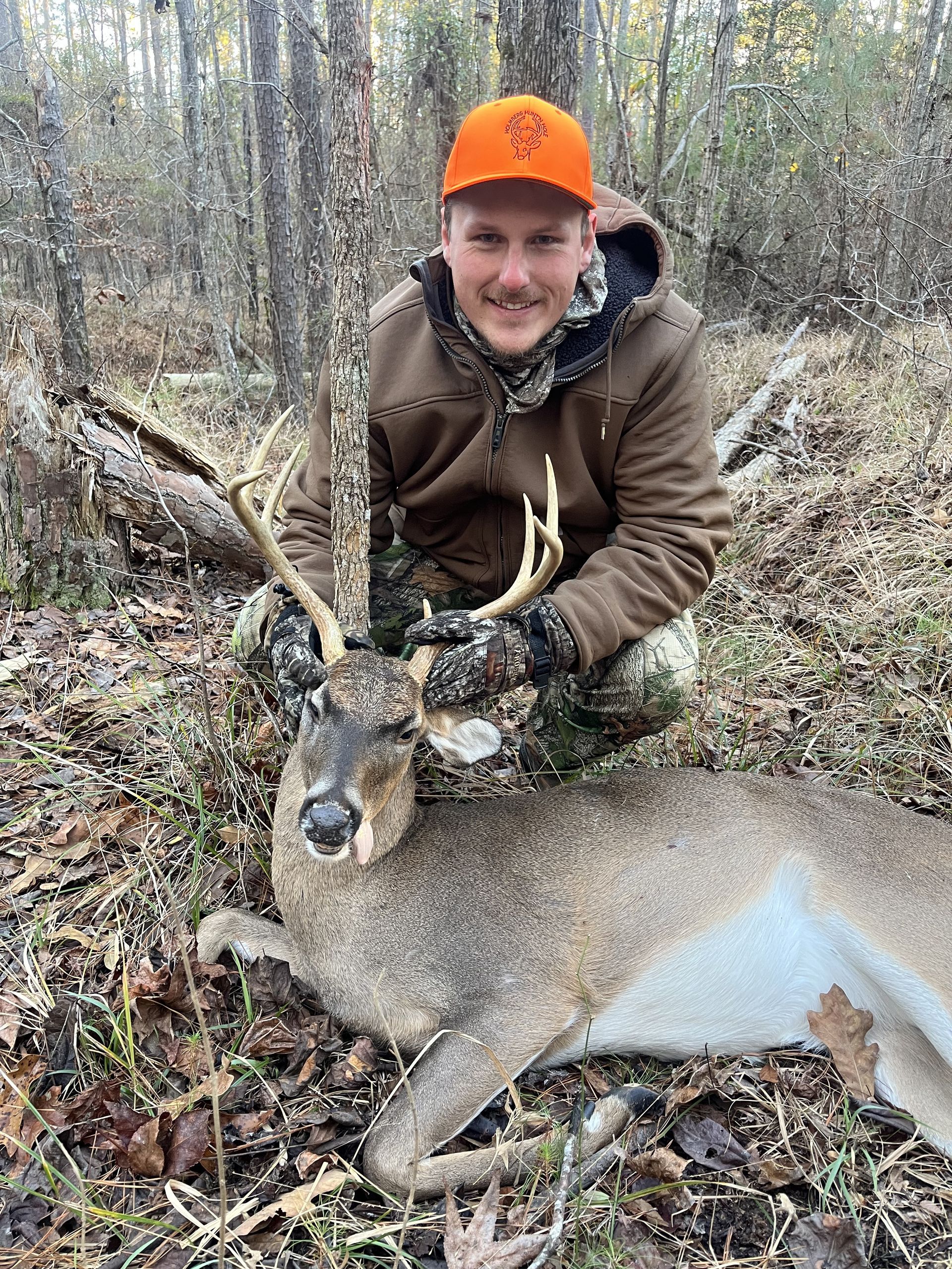 A smiling person kneels next to a harvested buck in a forest, wearing an orange hat and camouflage gear.