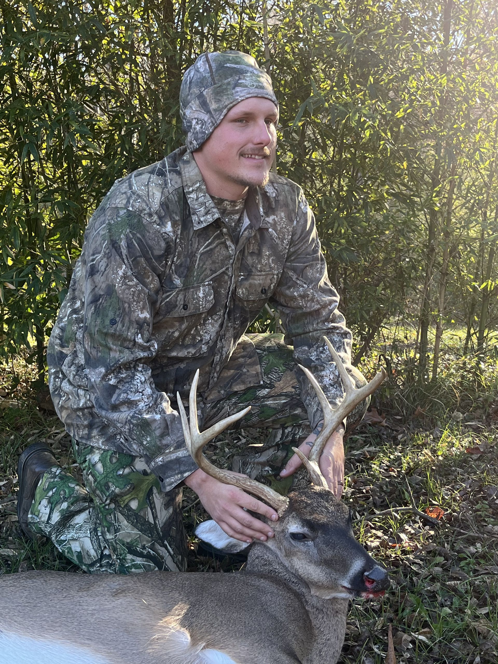 A man in camouflage kneels beside a harvested buck, smiling. The setting appears to be a wooded area with sunlight.