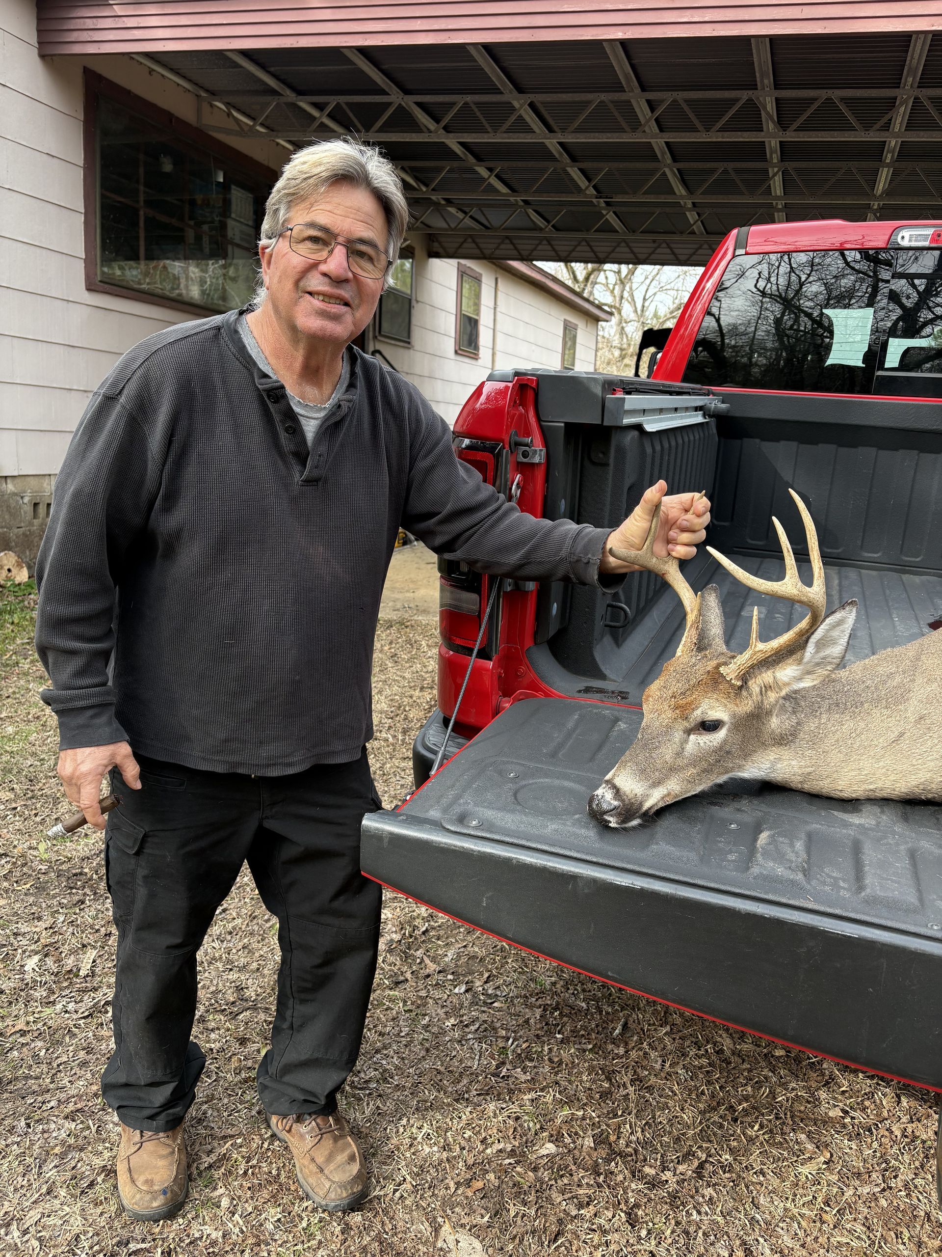 Man in a gray shirt stands next to a red truck, holding a deer's antlers in its bed. He is smiling in an outdoor setting.