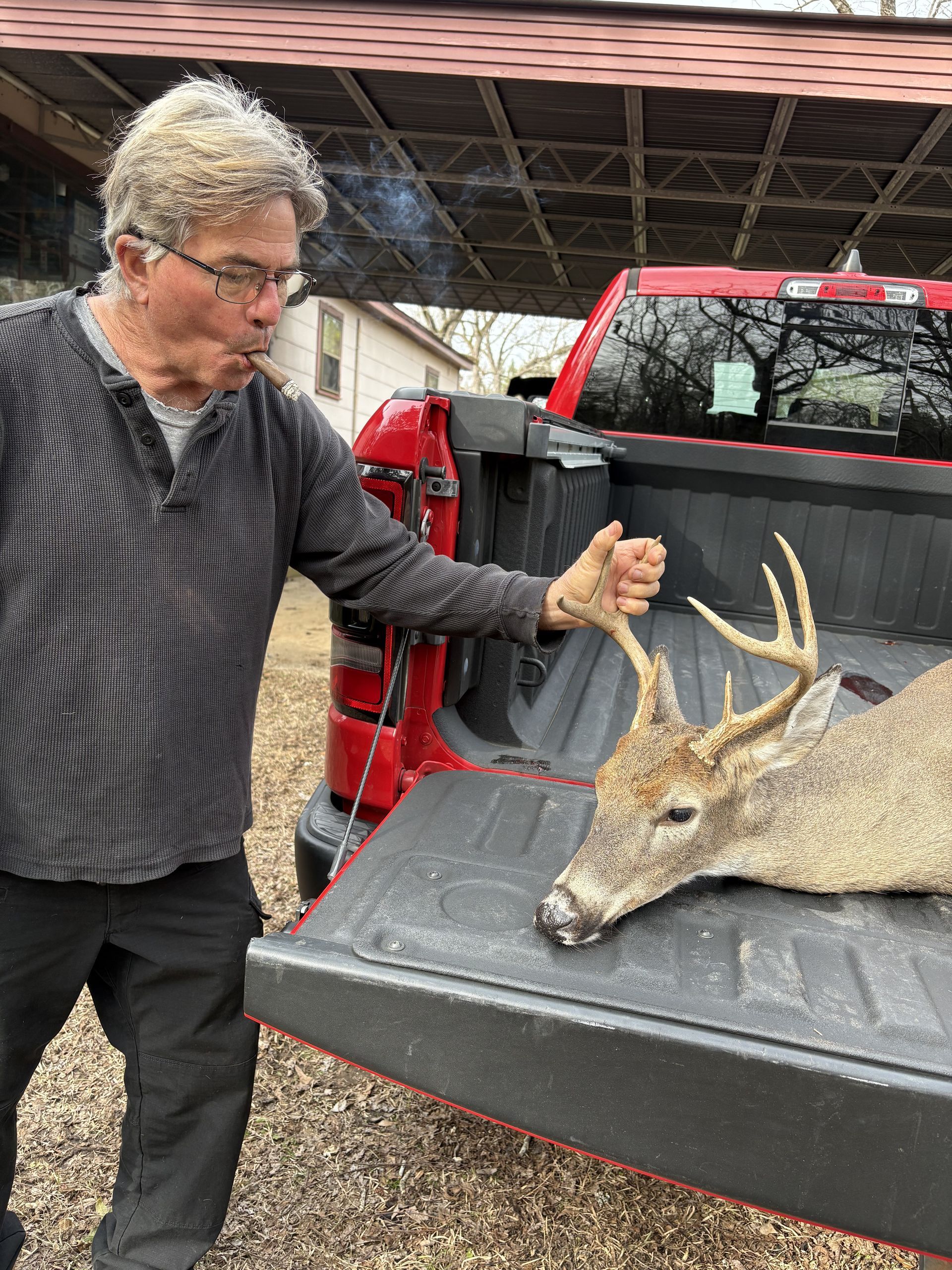Man exhales smoke, pointing at a dead buck in a red truck bed. Outdoors, day.