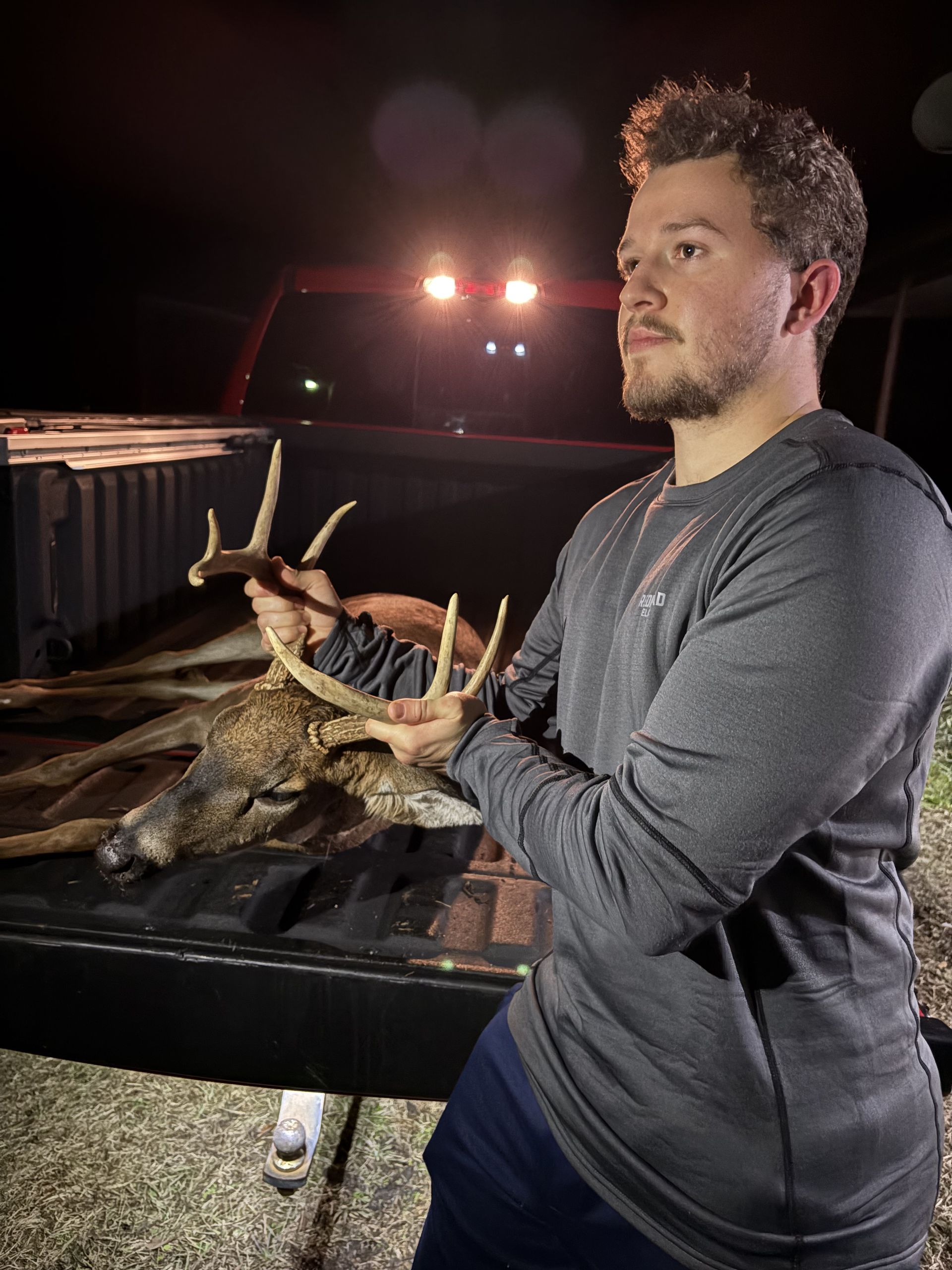 Man holding deer antlers in a truck bed at night, lit by truck lights. He's wearing a gray long-sleeve shirt, looking off to the side.