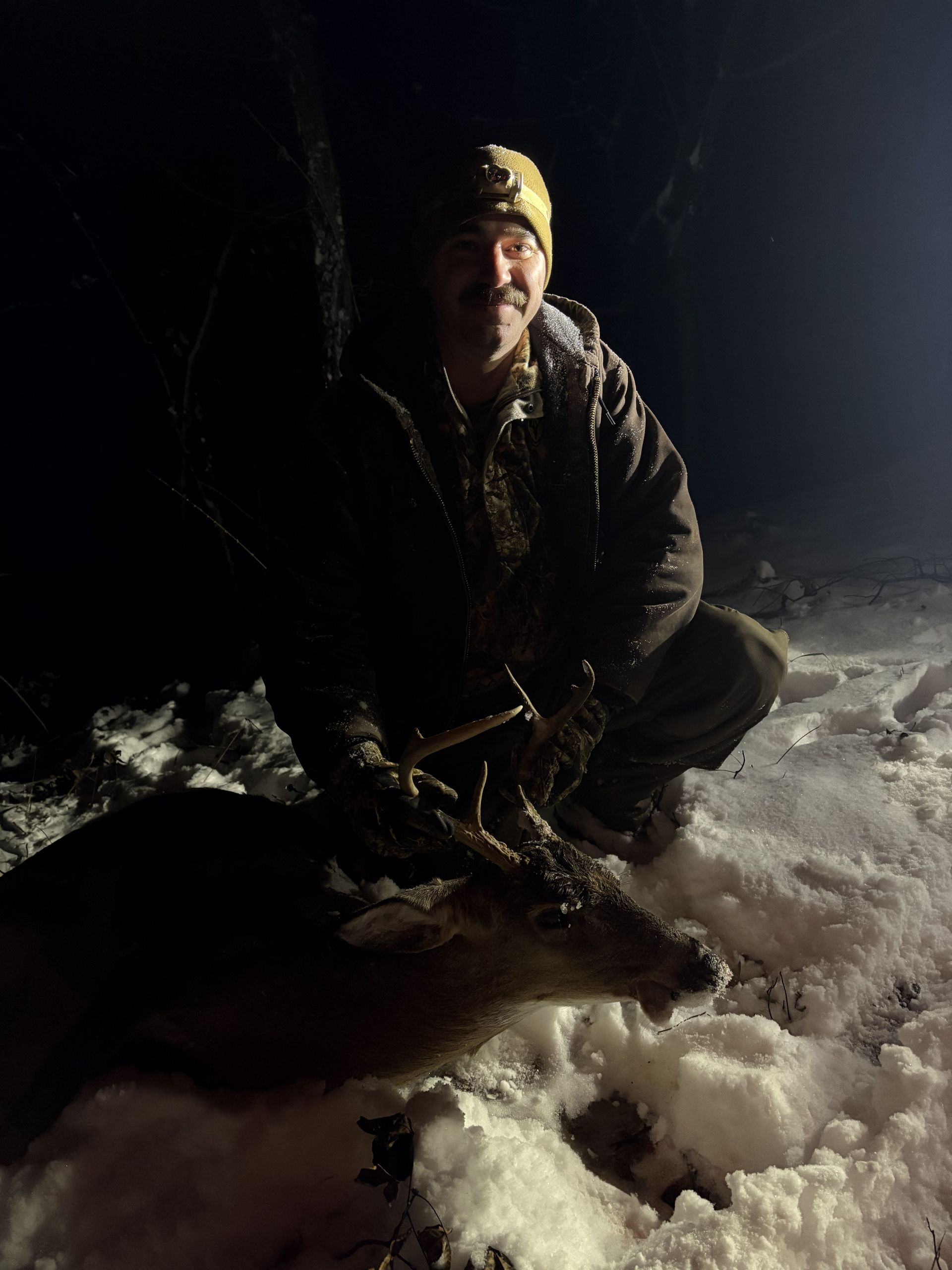 A man kneels in snow next to a deer he has hunted, illuminated by a flashlight at night. He is smiling.