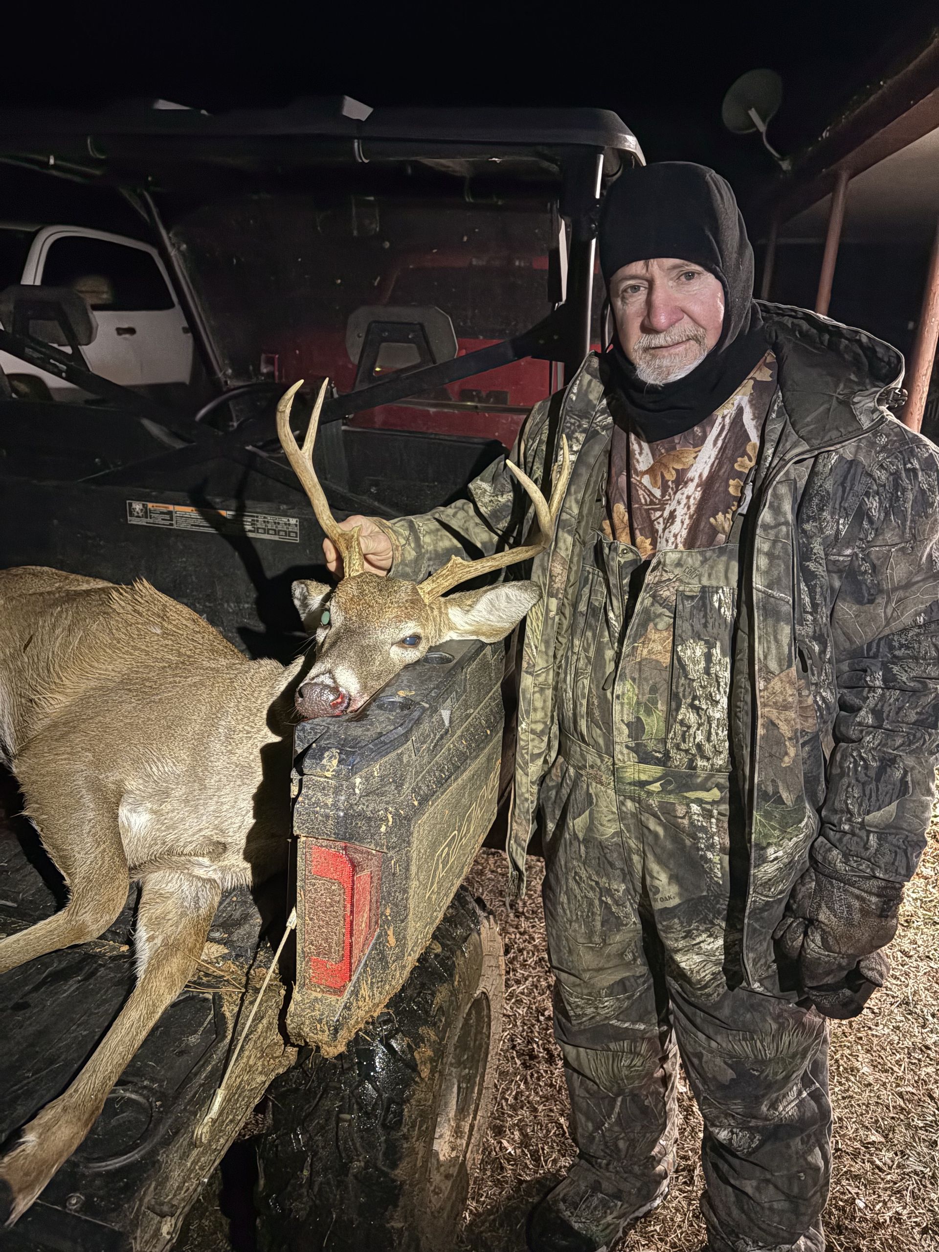 Man in camouflage stands next to a deer in the back of a truck at night. He wears a hat and looks at the camera.