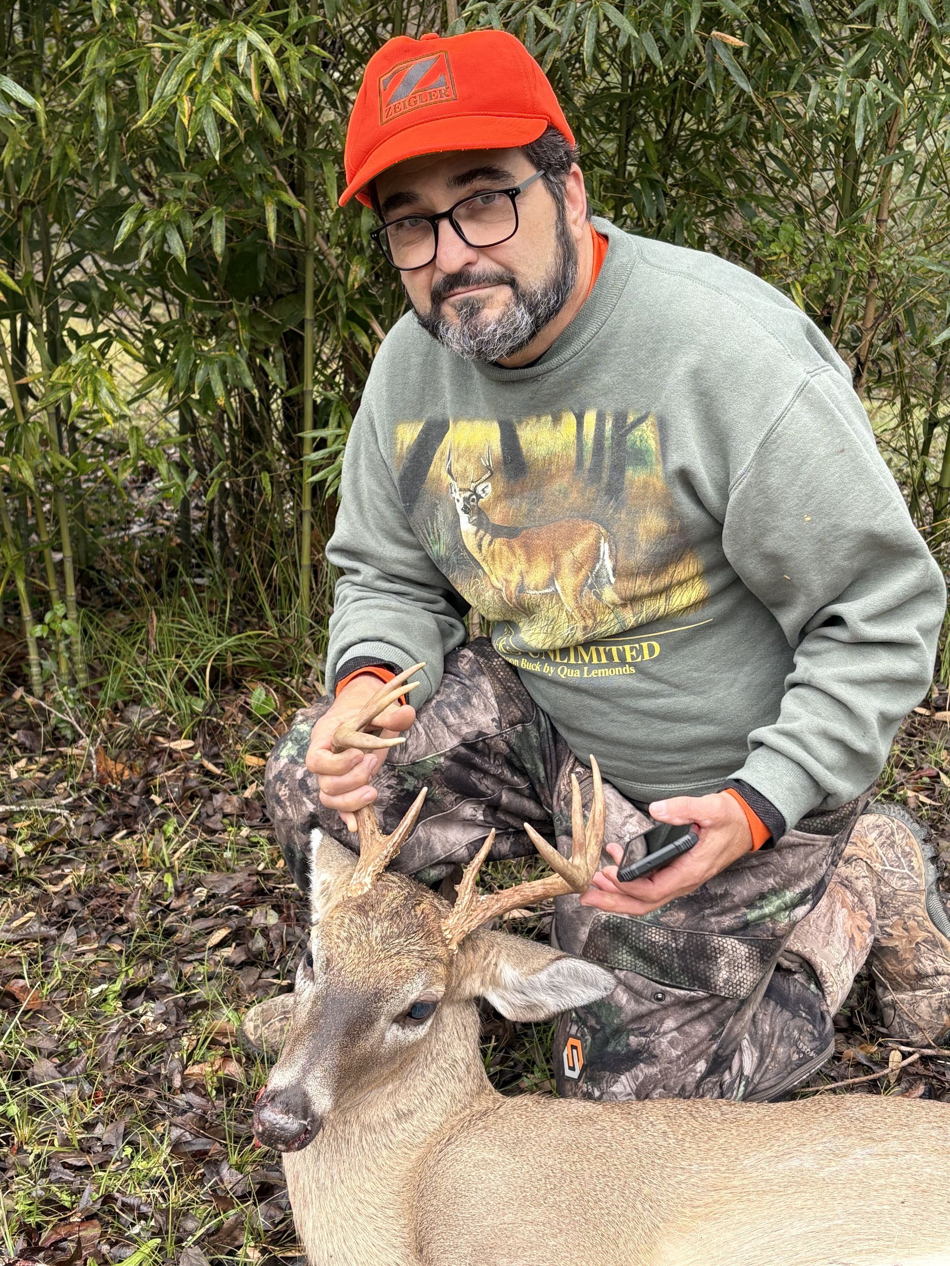 A man in hunting gear kneels beside a harvested buck with antlers. He wears an orange cap and smiles, set in a wooded area.