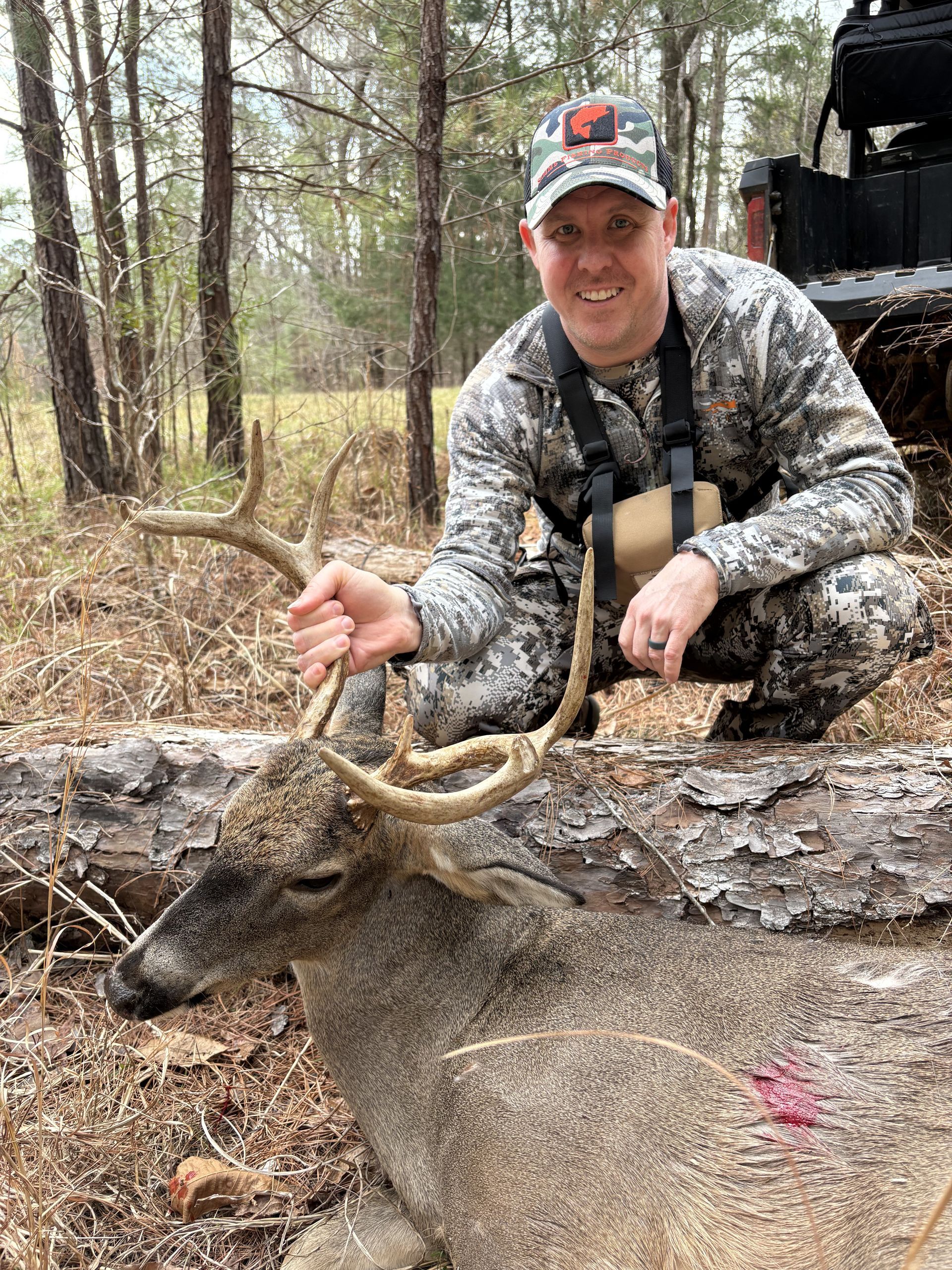 Man in camo kneels beside a harvested deer in a wooded area, smiling and holding the antlers.