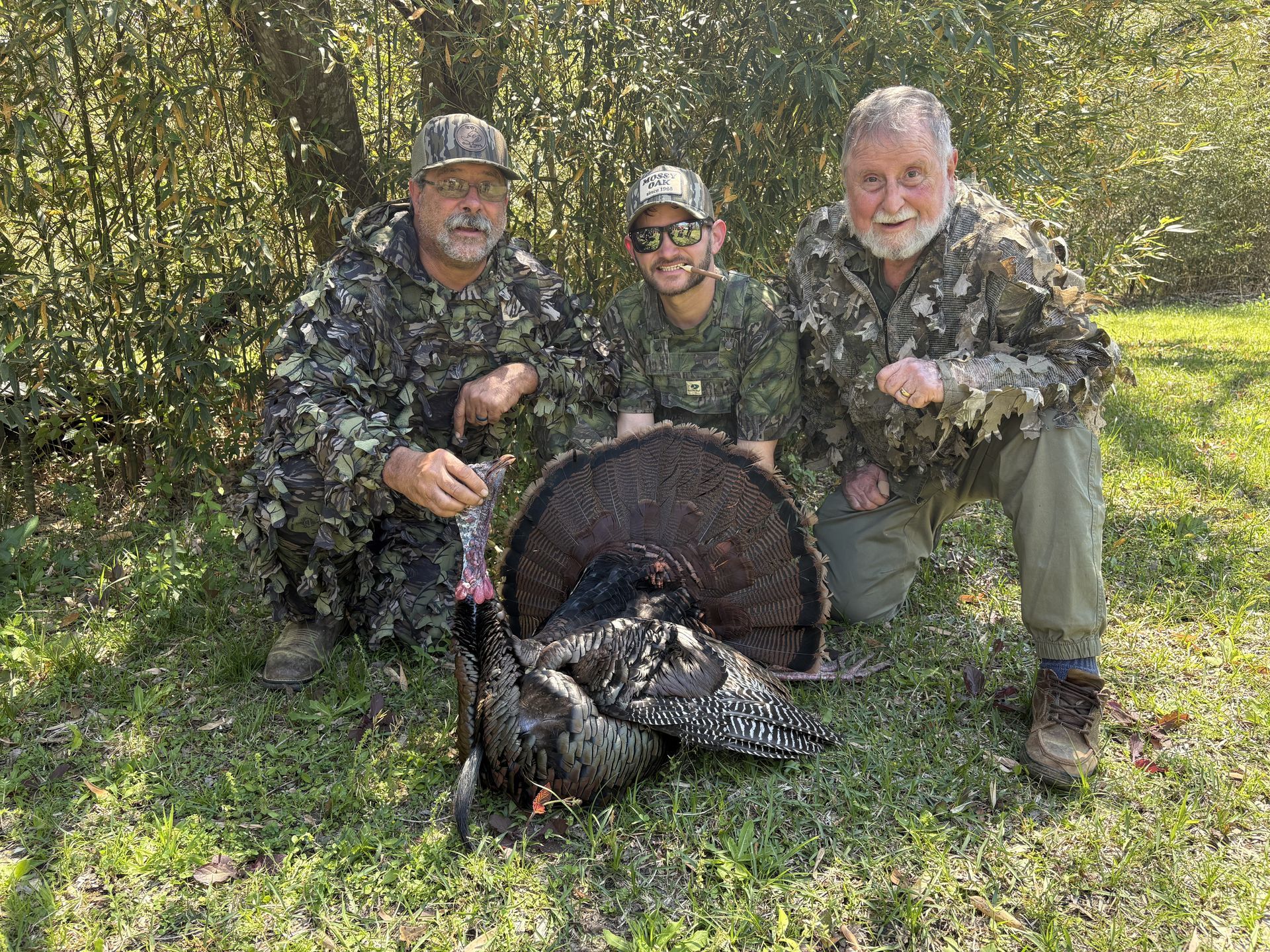Three men in camouflage pose with a dead turkey outdoors. They kneel on grass in a sunny wooded area, smiling.