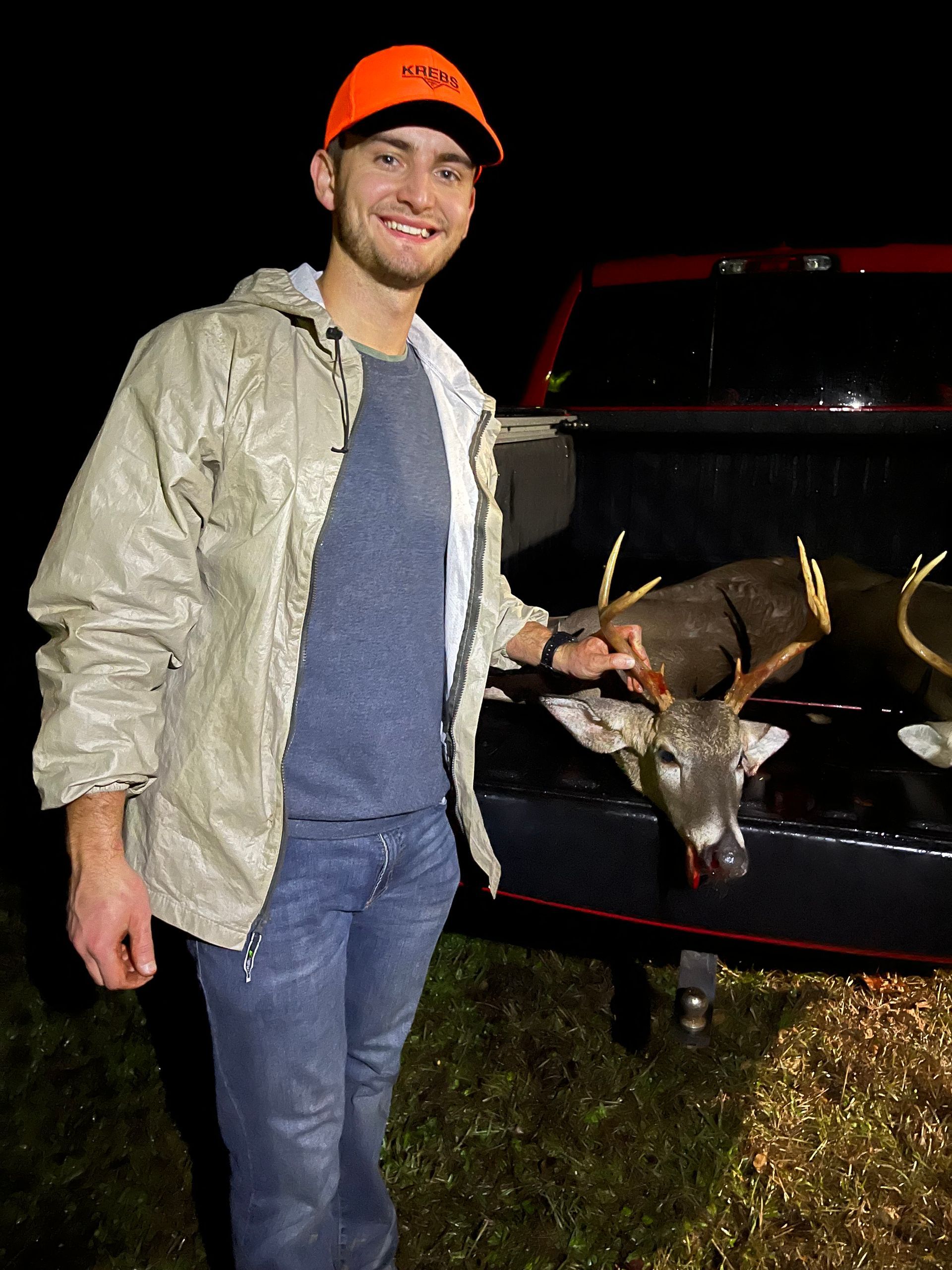Man wearing orange hat and tan jacket stands beside a truck bed containing two deer carcasses. He smiles at the camera.