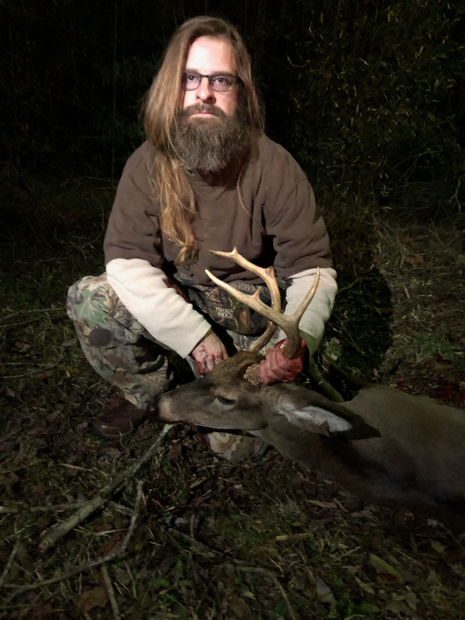 Man with long hair and beard kneels beside a harvested buck in a forest at night. He's wearing glasses, a brown shirt, and camouflage pants.