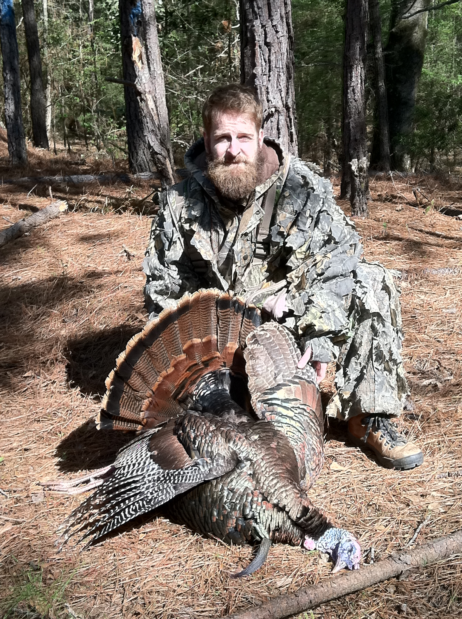 Man in camouflage kneeling with a dead turkey in a wooded area. The man has a long beard.