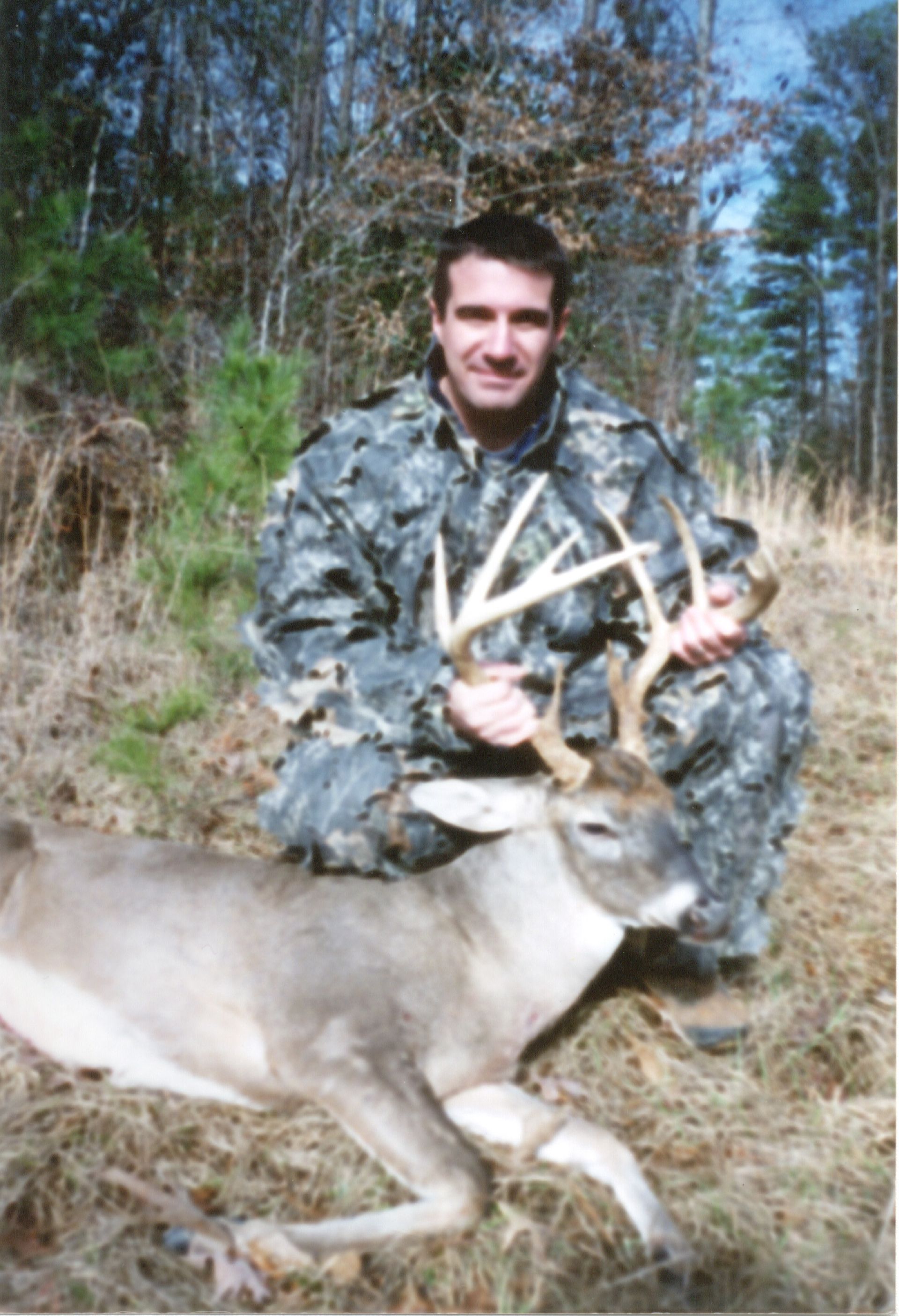 A man in camouflage kneels beside a dead deer in a wooded area, holding the deer's large antlers and smiling.