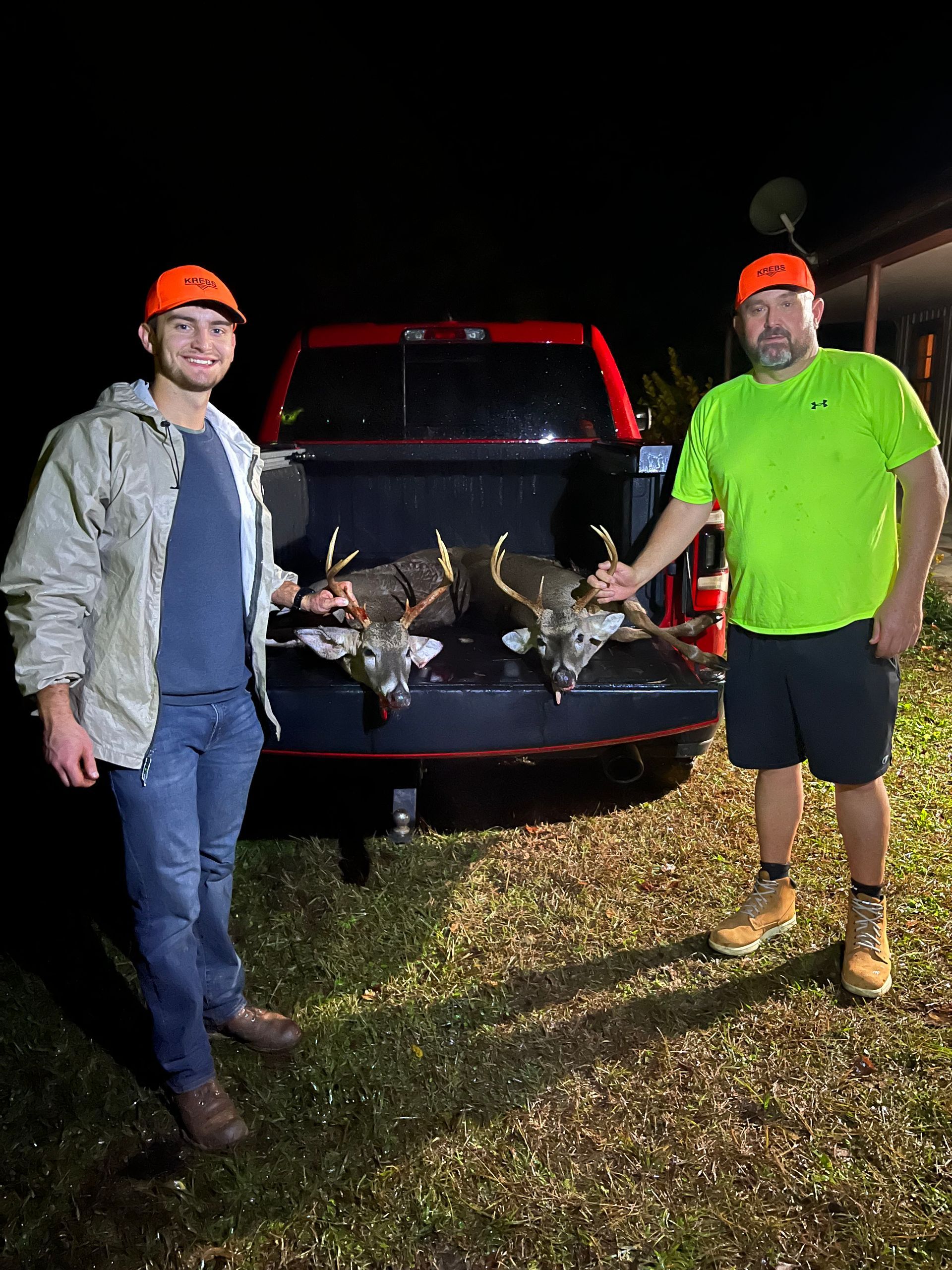 Two men stand beside the bed of a truck, each holding a deer head. They wear orange hats; it's nighttime.
