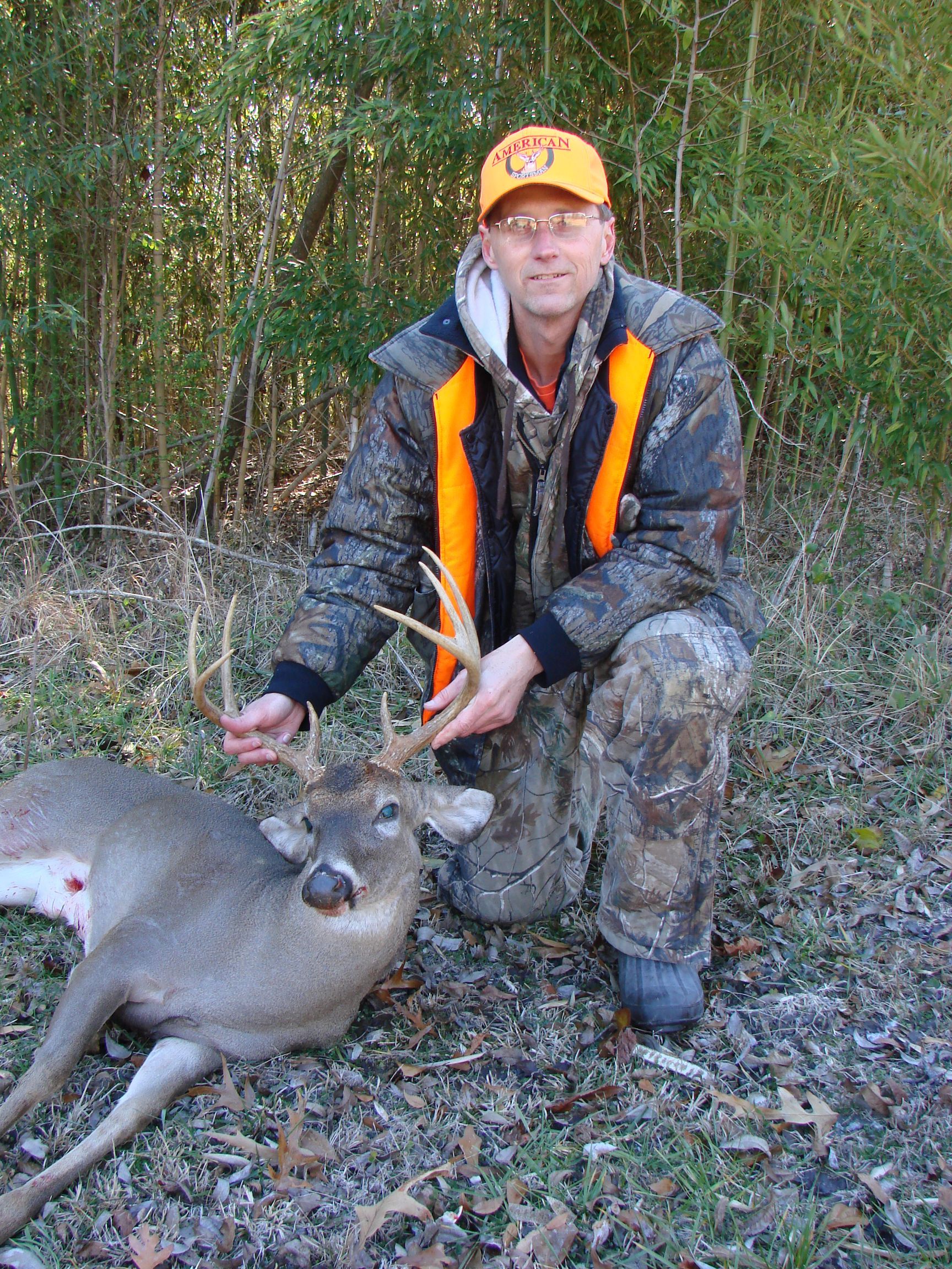 Man in camouflage kneels next to a dead deer in a forest. He wears an orange vest and hat, holding the deer's antlers.