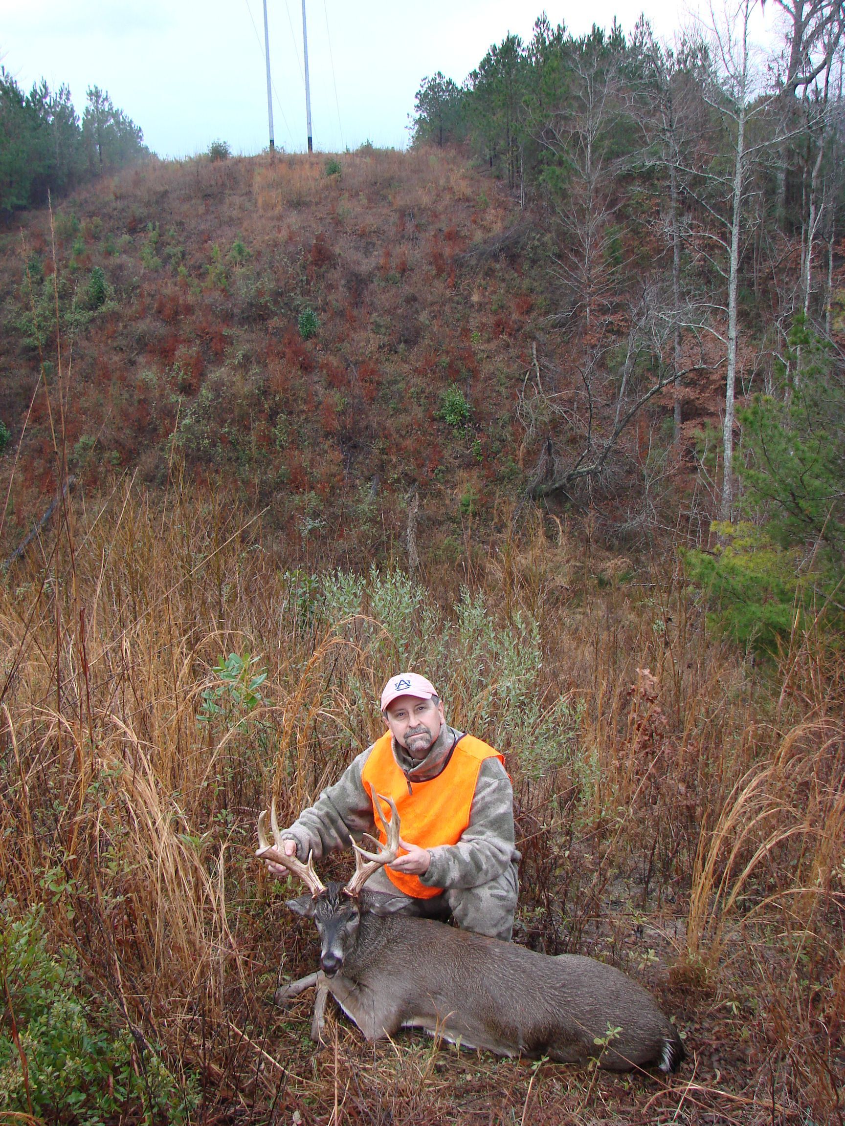 Hunter in orange vest kneels beside a deer he has shot, with tall brown grass and a hillside in the background.