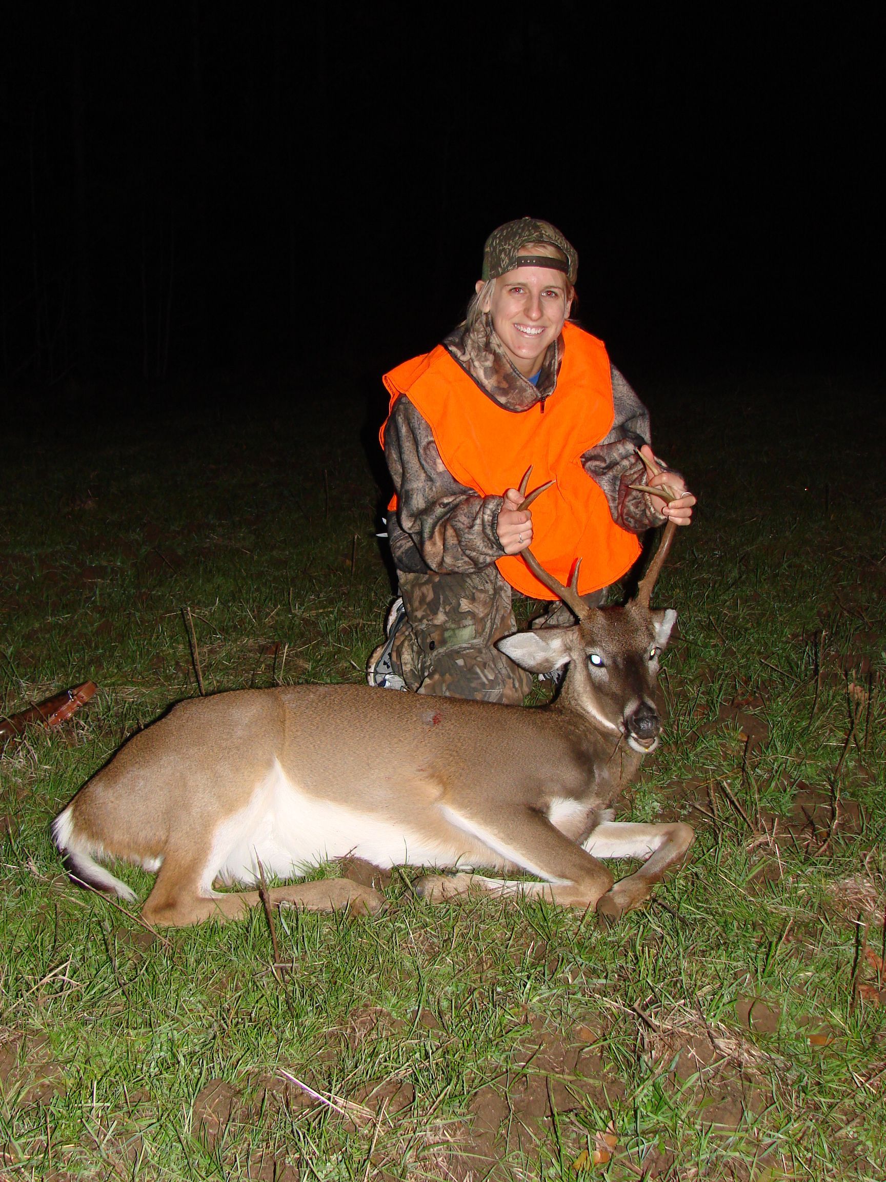 Woman in camouflage and orange vest kneels with a deer on grassy ground at night, holding its antlers and smiling.