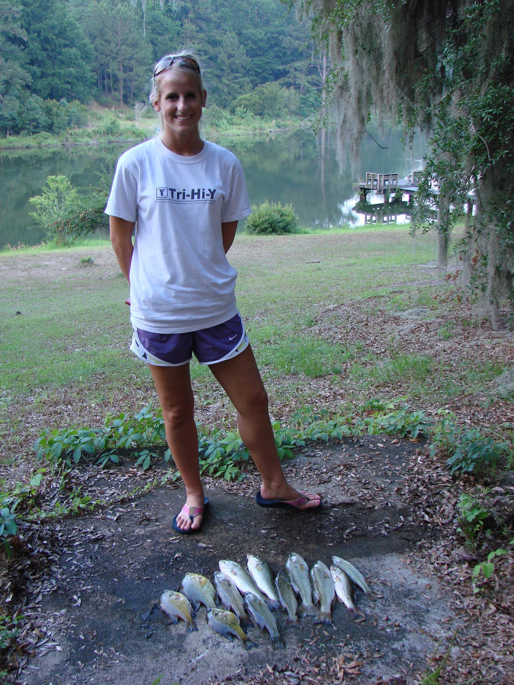 Woman standing near a lake, smiling and showing off a pile of freshly caught fish.
