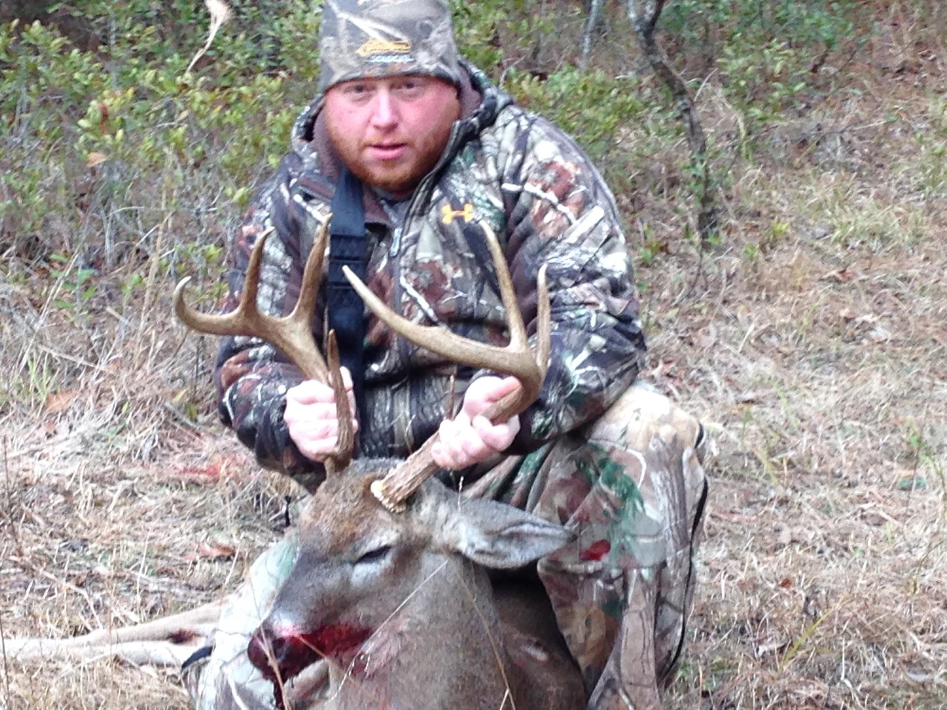 Man in camouflage with a deer he hunted. He kneels beside the deer, holding its antlers in a wooded setting.