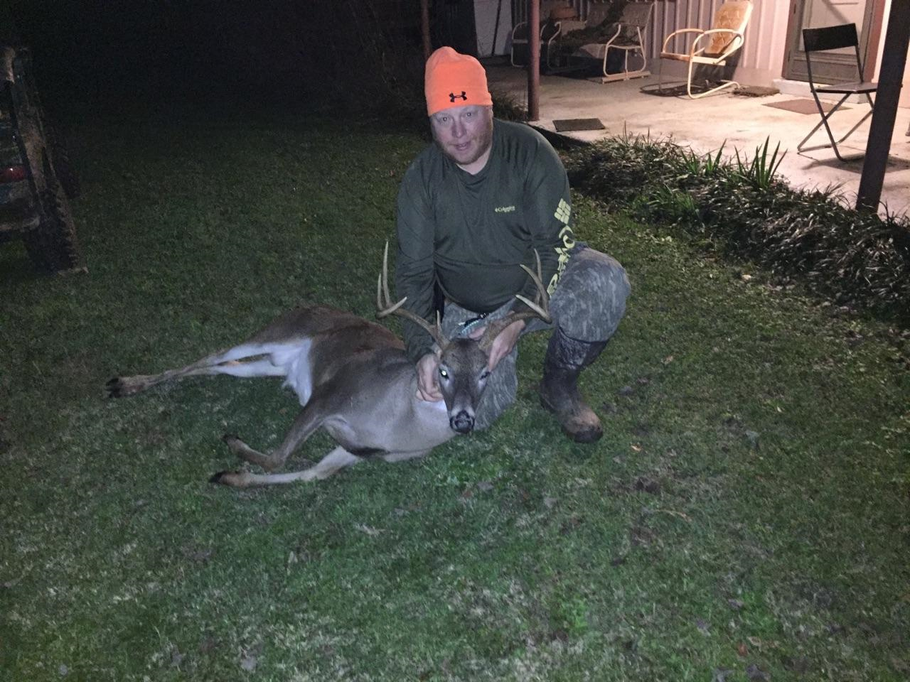 A man kneels next to a dead deer on a lawn at night. He wears an orange hat and holds the deer's antlers.
