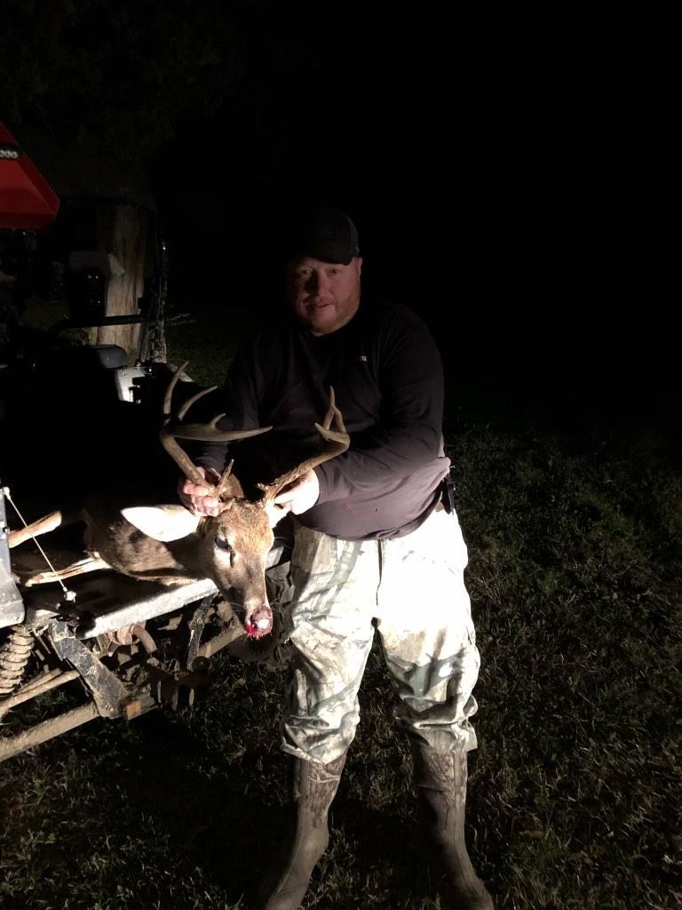 A man in hunting gear stands next to an ATV, holding a deer he has apparently hunted. It's night, and the scene is illuminated.
