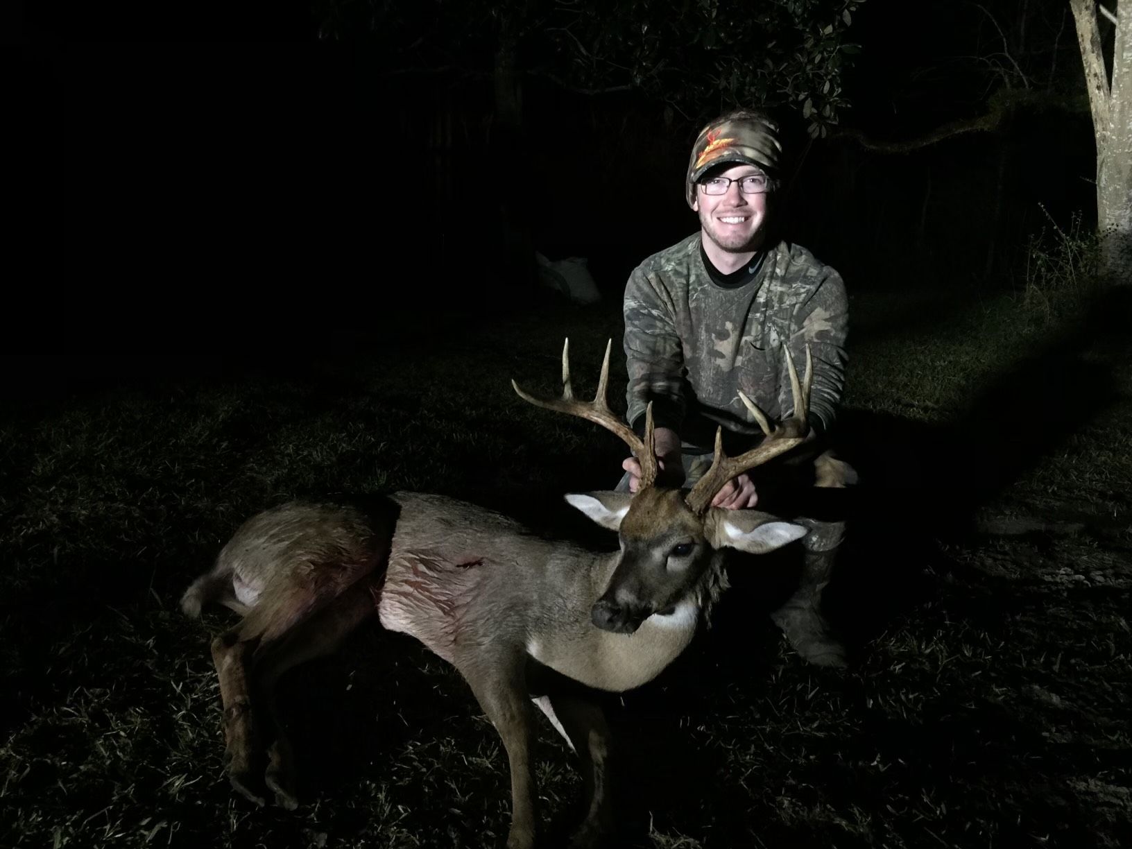 A man in camouflage smiles, kneeling next to a deer he has killed. The scene is outdoors and dark.