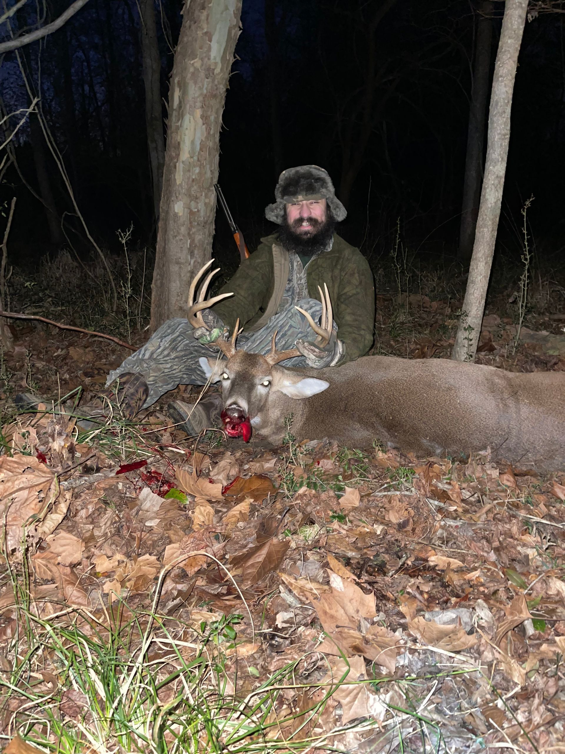 Man kneels beside two deer he hunted in a wooded area at night. The deer have antlers and blood near their mouths.