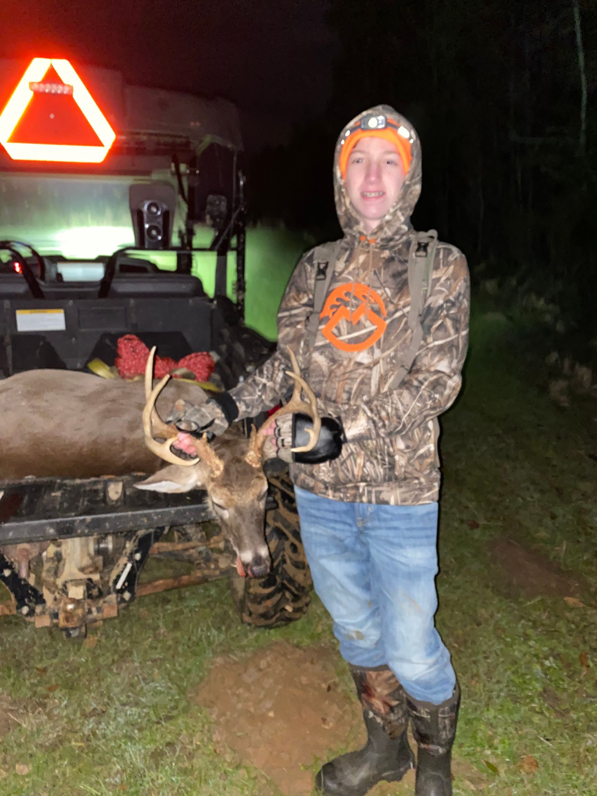 A person in camo clothing poses with a buck in the back of a utility vehicle at night. The deer has antlers.